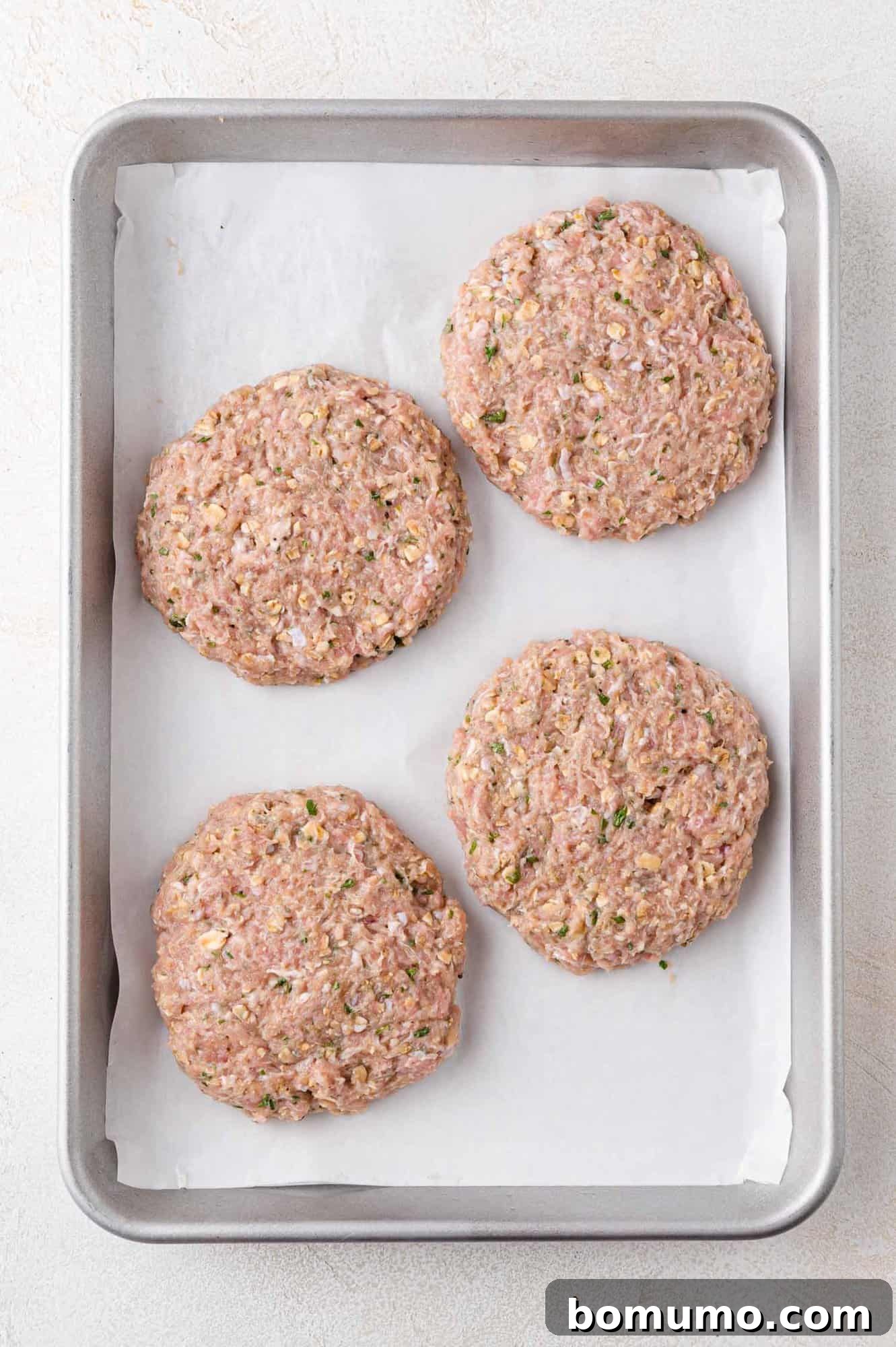 Four perfectly formed raw turkey burger patties, seasoned with visible herbs, carefully arranged on a parchment-lined baking sheet, awaiting their cooking method.