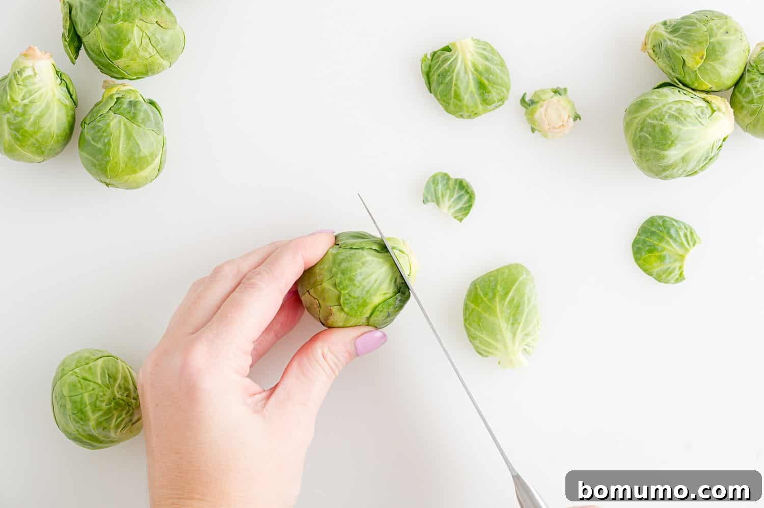 A hand using a knife to trim the stem off a whole Brussels sprout, demonstrating the initial prep step.