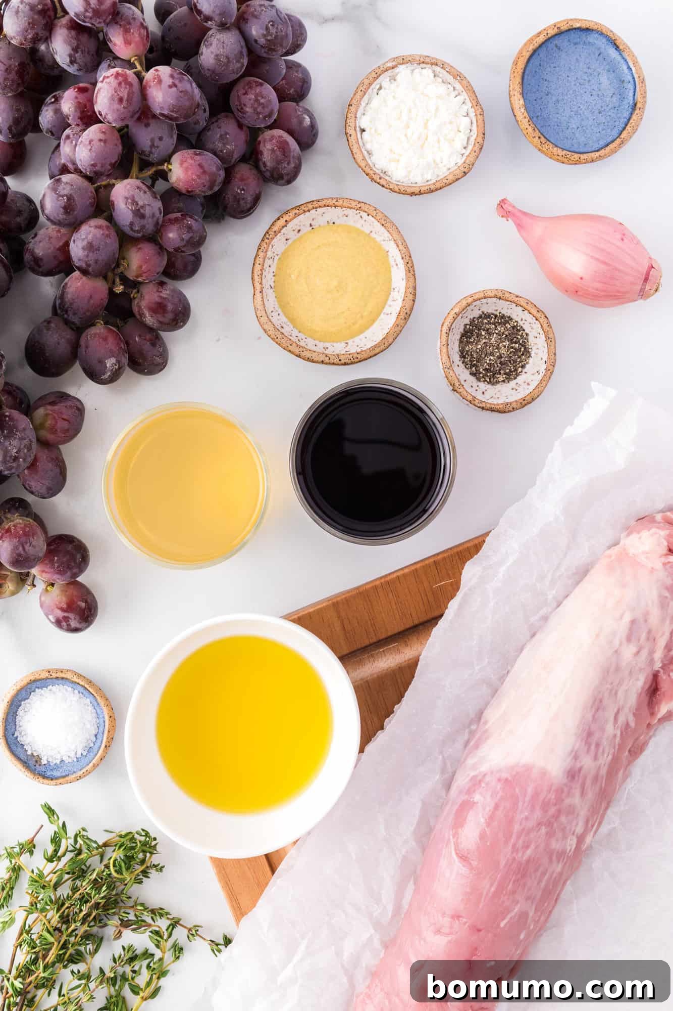 A vibrant display of fresh ingredients for the recipe, including plump red grapes, a trimmed pork tenderloin, shallots, and various herbs and spices.