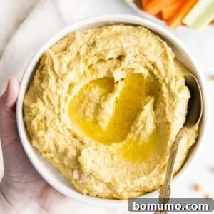 Hummus in a white bowl with a spoon, topped with olive oil and a sprinkle of paprika, ready to be enjoyed.