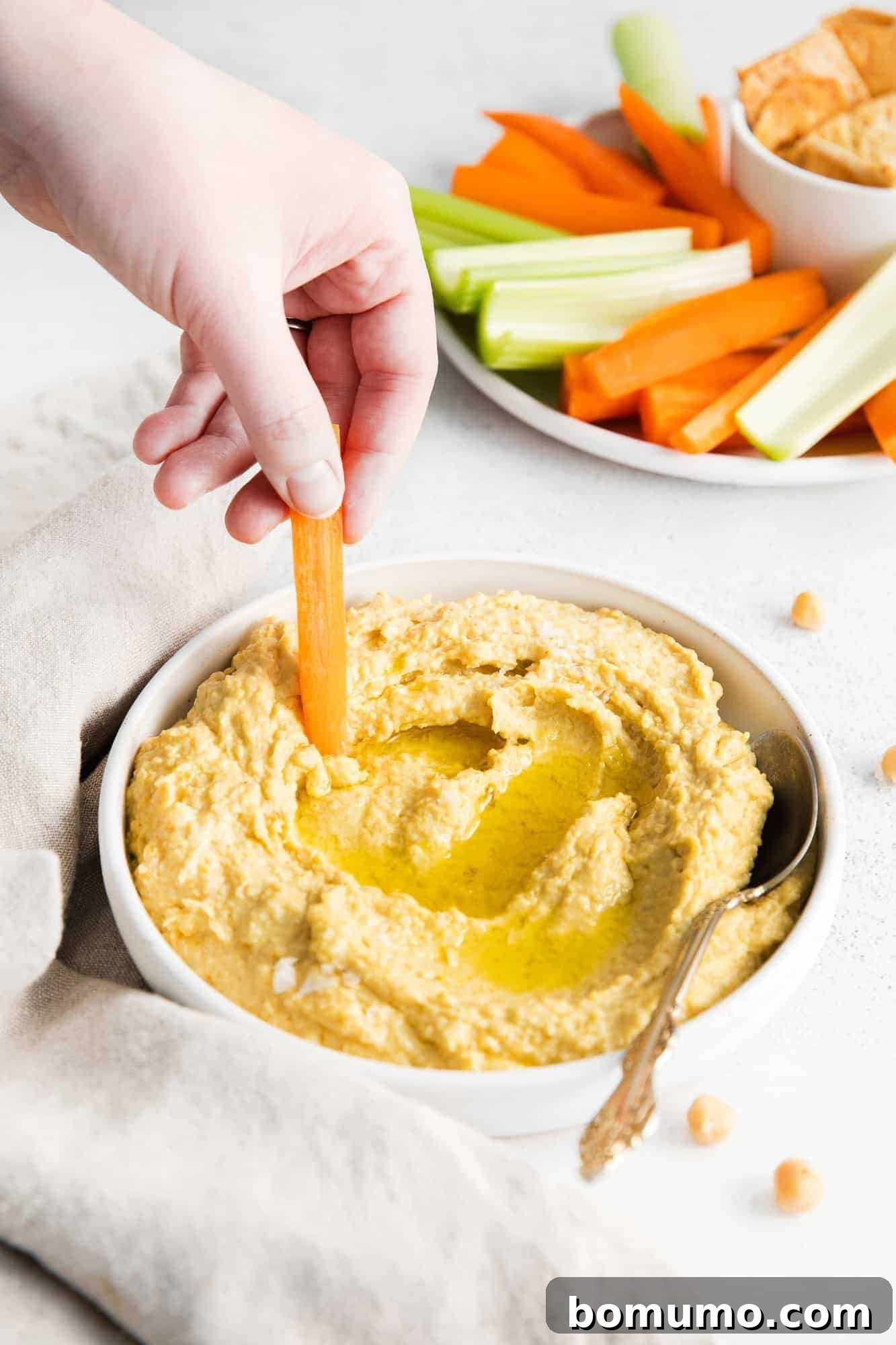 A hand dipping a vibrant carrot stick into a bowl of perfectly smooth homemade hummus, showcasing its creamy texture and inviting appeal.