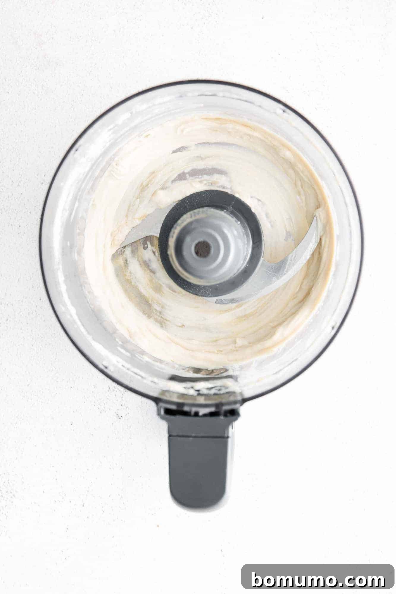 Overhead view of tahini, lemon juice, and water being processed into a light, airy mixture in a food processor, the first step to creamy hummus.