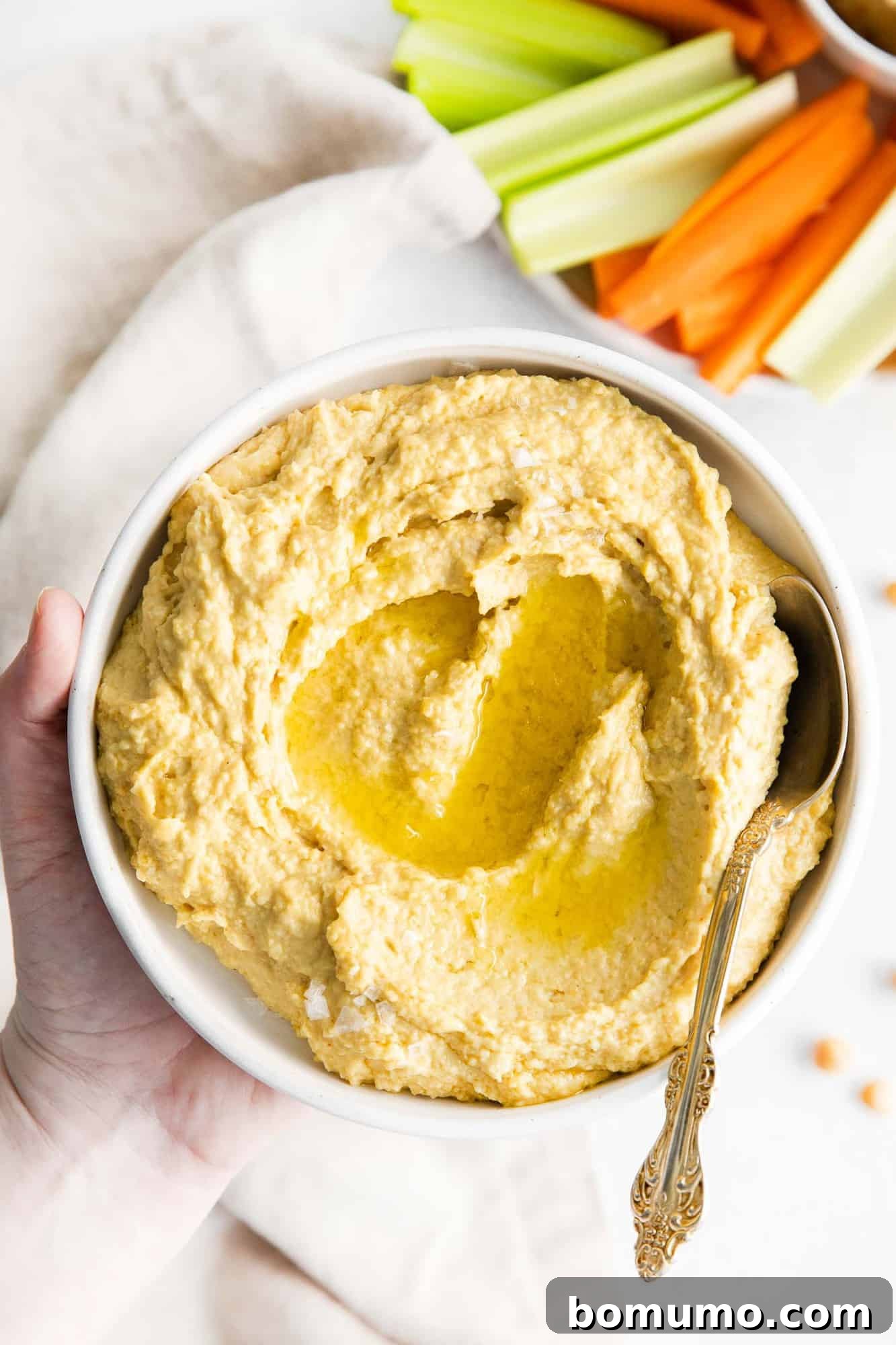 A hand holding a bowl of hummus, garnished with olive oil and spices, with a side of carrot and celery sticks for dipping.