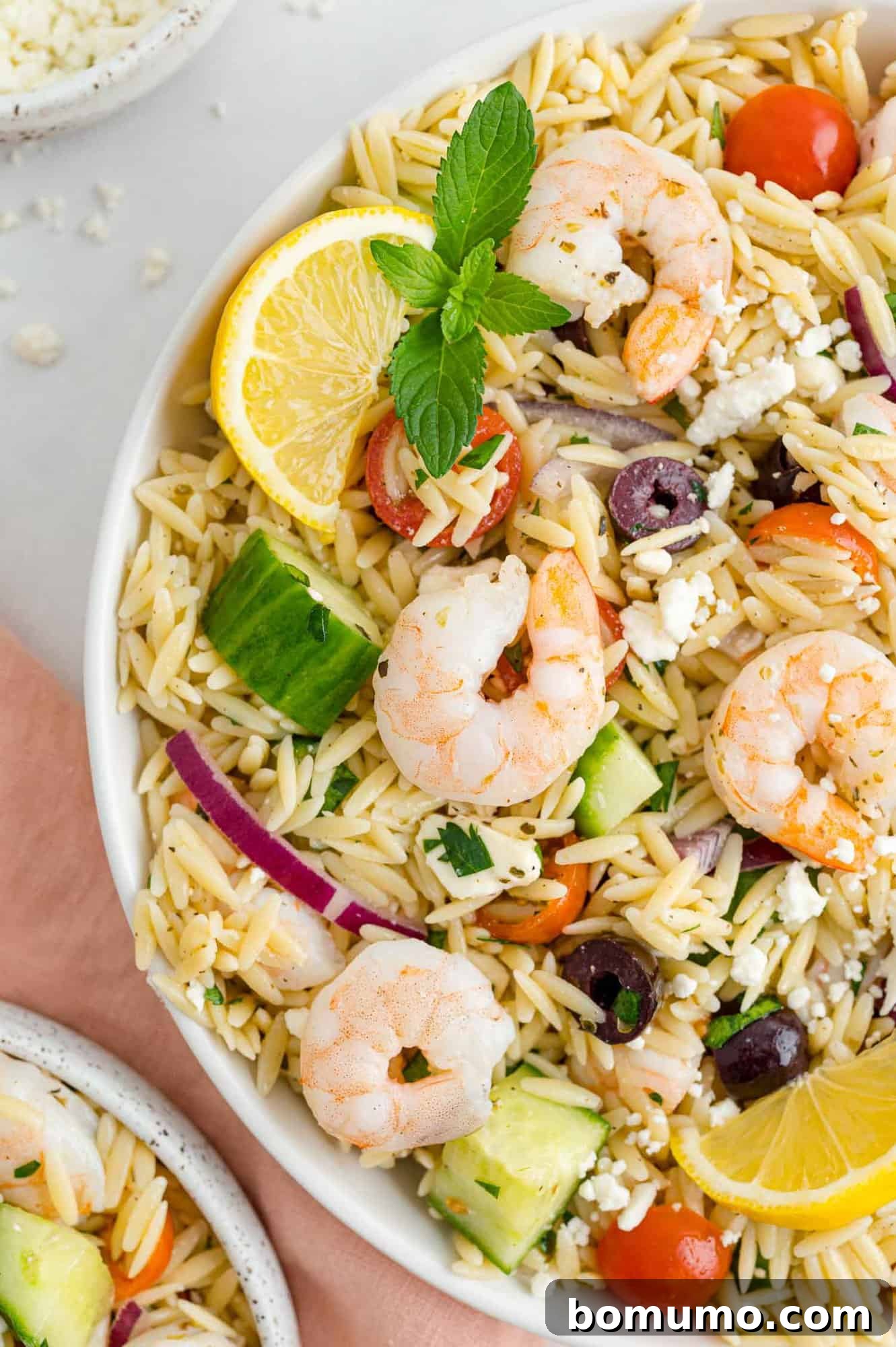 A vibrant Greek orzo salad featuring tender shrimp, bright lemon wedges, and fresh mint leaves, presented in a stylish bowl.