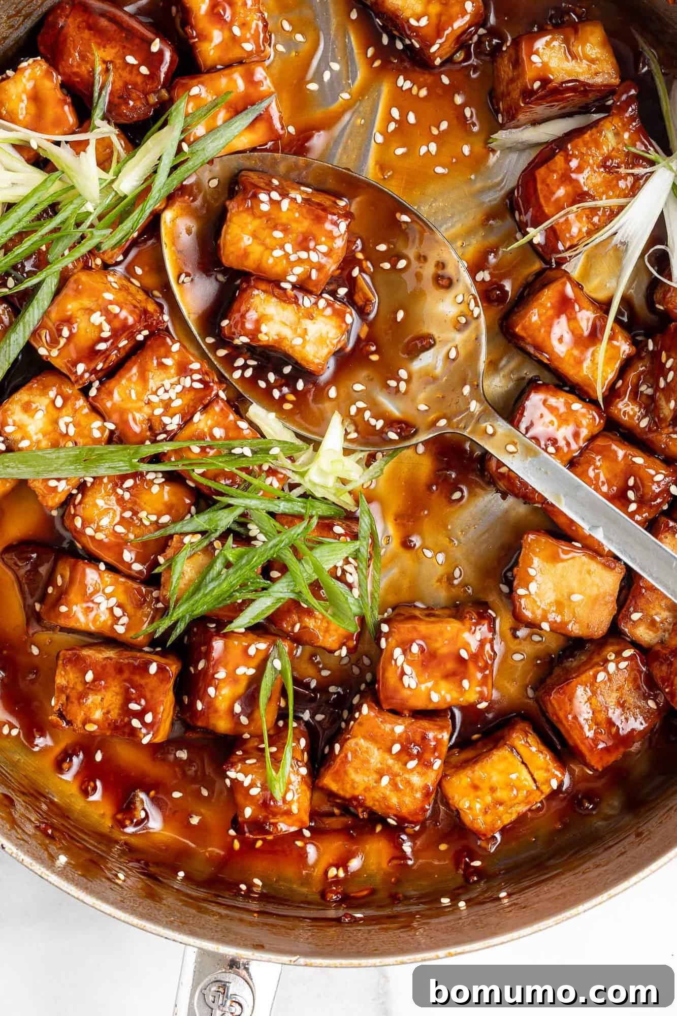 Overhead view of sticky sesame tofu in a frying pan with vibrant green onions scattered across.