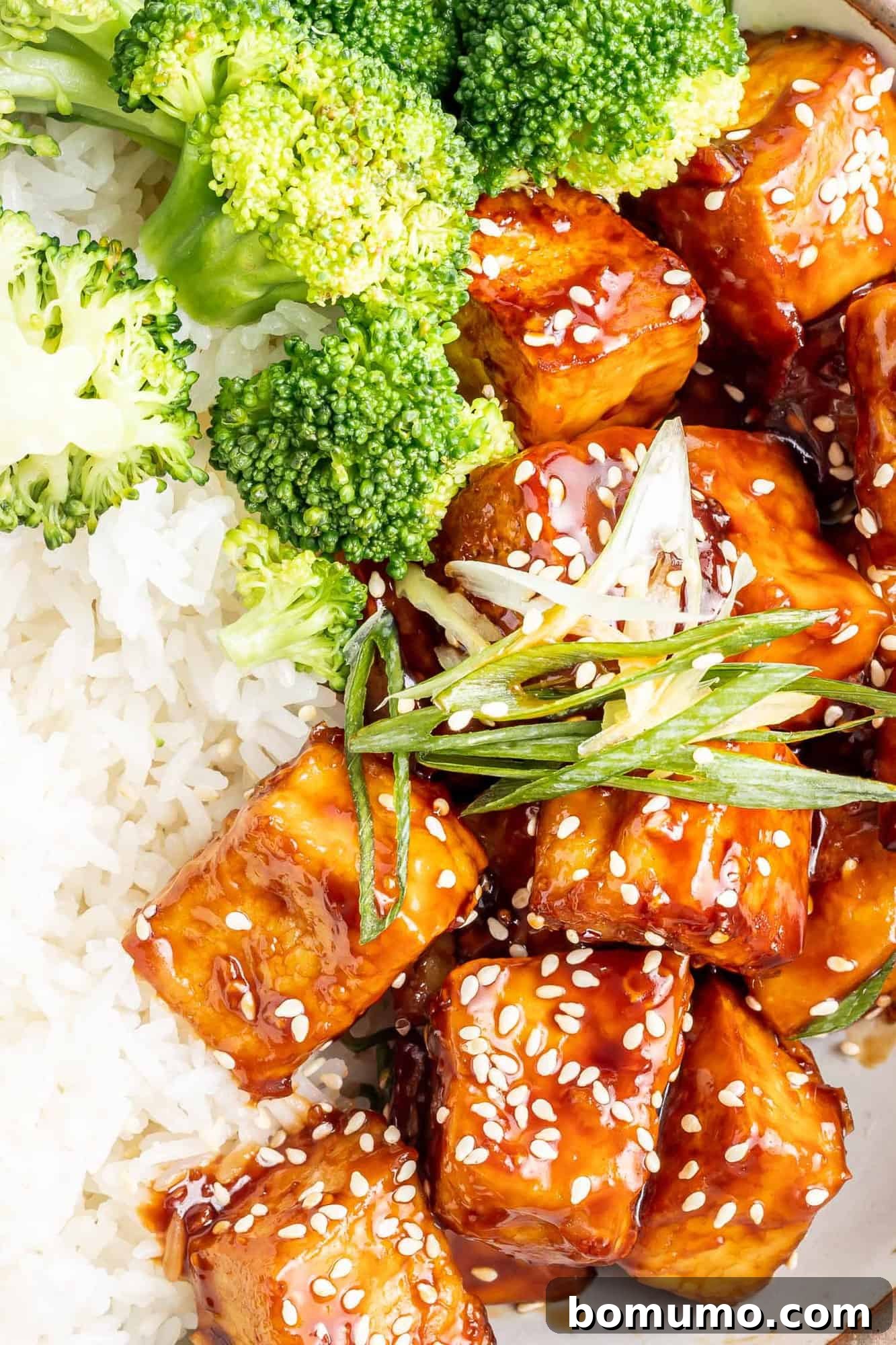 Close up of perfectly cooked sesame tofu served alongside a bed of white rice and fresh broccoli florets, lightly sprinkled with sesame seeds for garnish.
