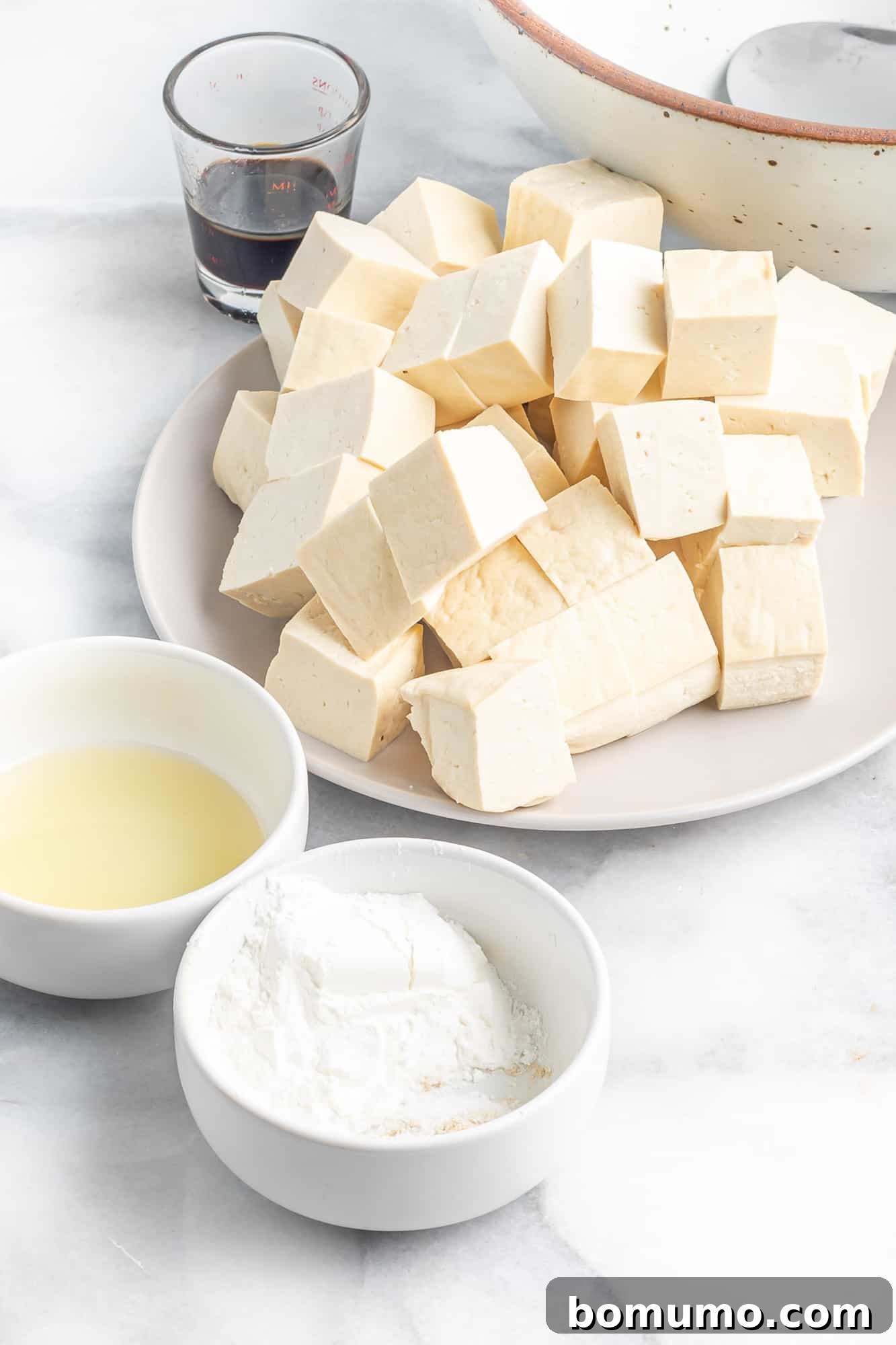 A selection of ingredients neatly arranged, including a block of tofu, a bowl of cornstarch, and a bottle of cooking oil, ready for use.
