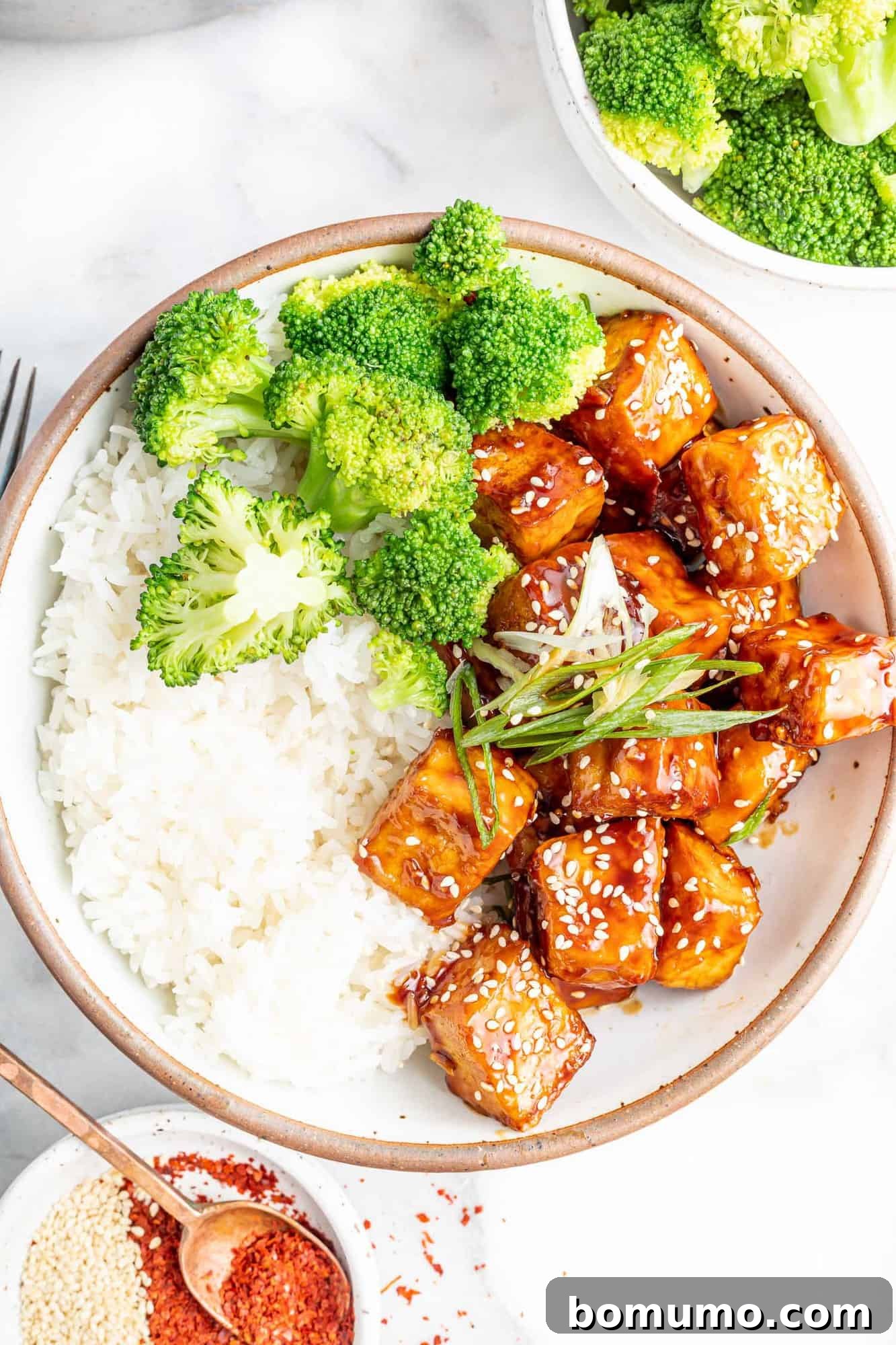 Overhead view of prepared sesame tofu in a bowl, accompanied by rice and broccoli, beautifully garnished with sesame seeds.