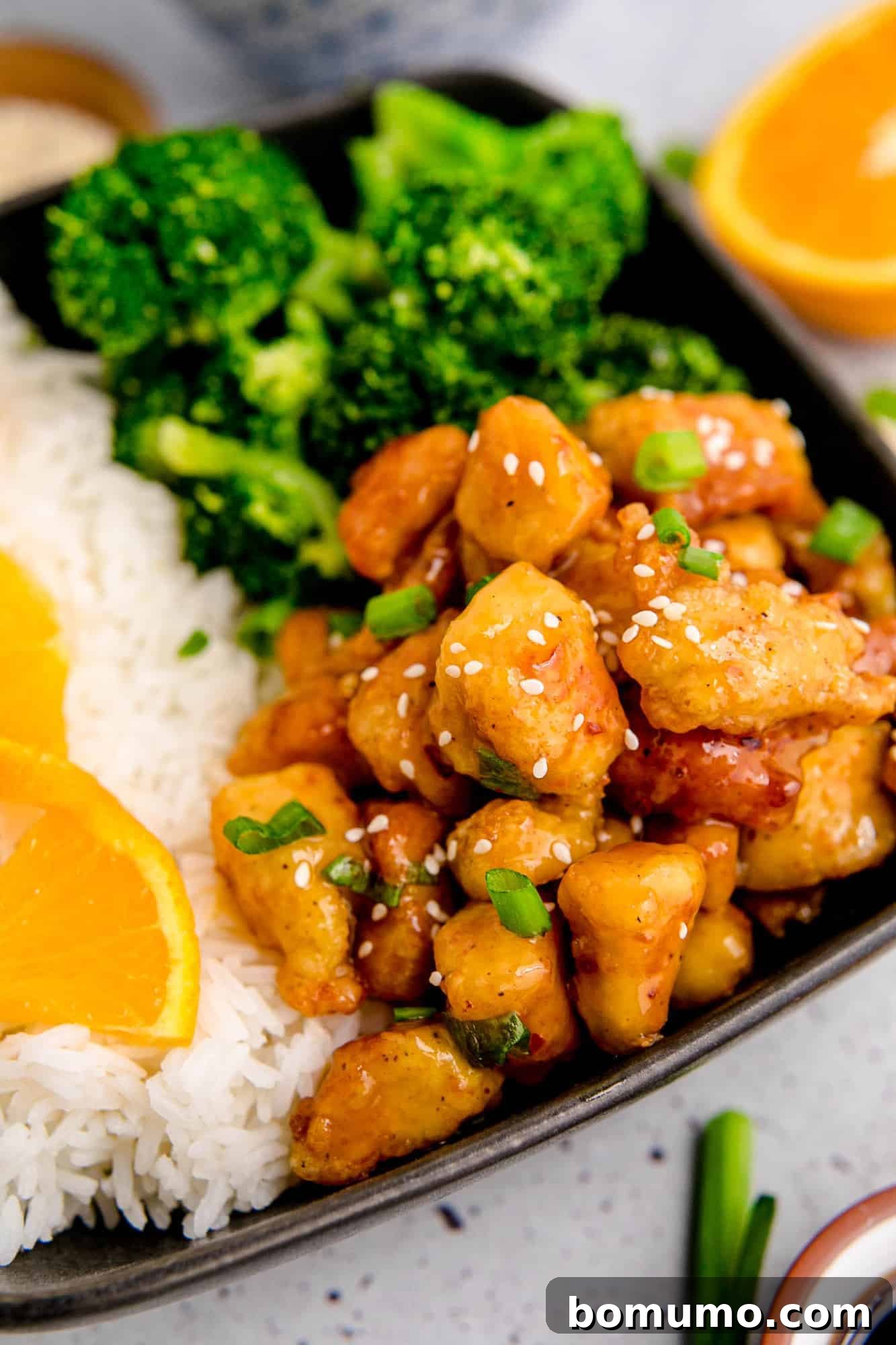 Homemade orange chicken on a black plate with rice and broccoli, garnished with sesame seeds.