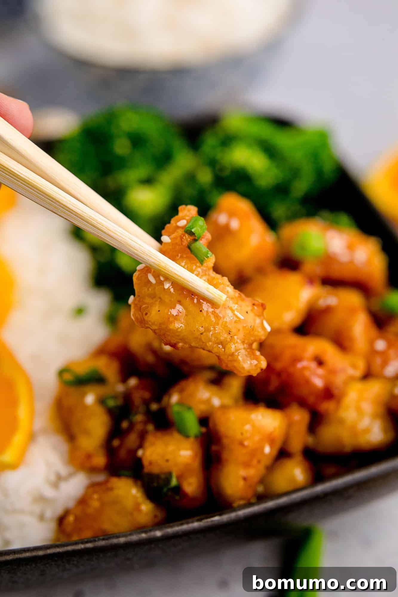 Crispy piece of glazed chicken held between chopsticks, with more orange chicken in the background.