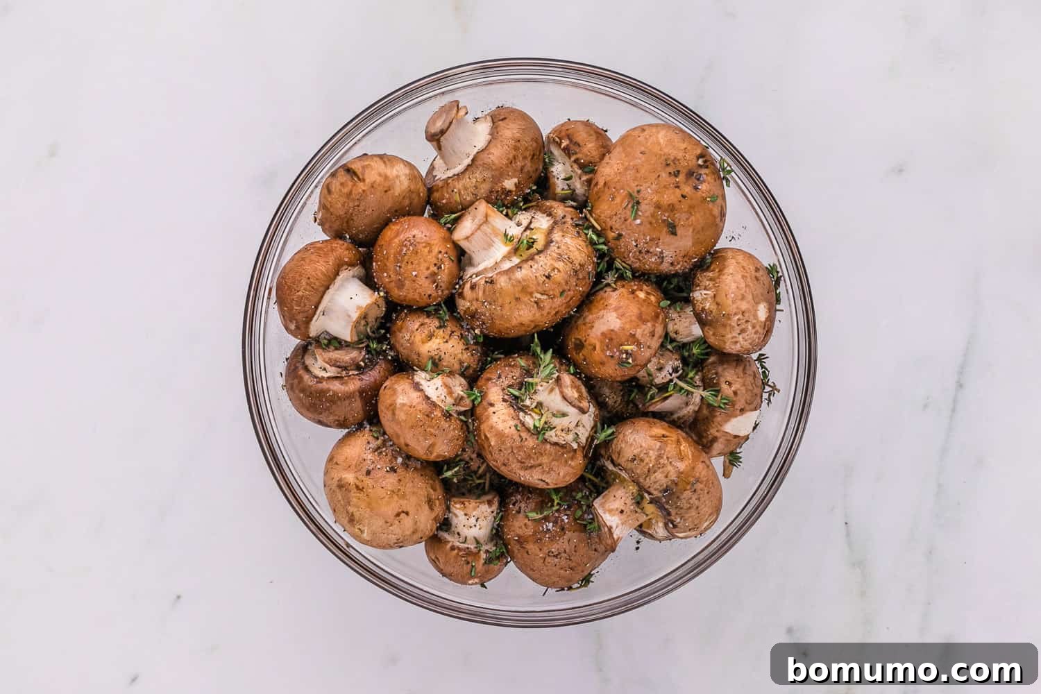 Umami Roasted Mushrooms 5 Uncooked mushrooms coated in a rich mixture of olive oil, balsamic vinegar, and seasonings in a clear glass bowl, ready to be spread onto a baking sheet.