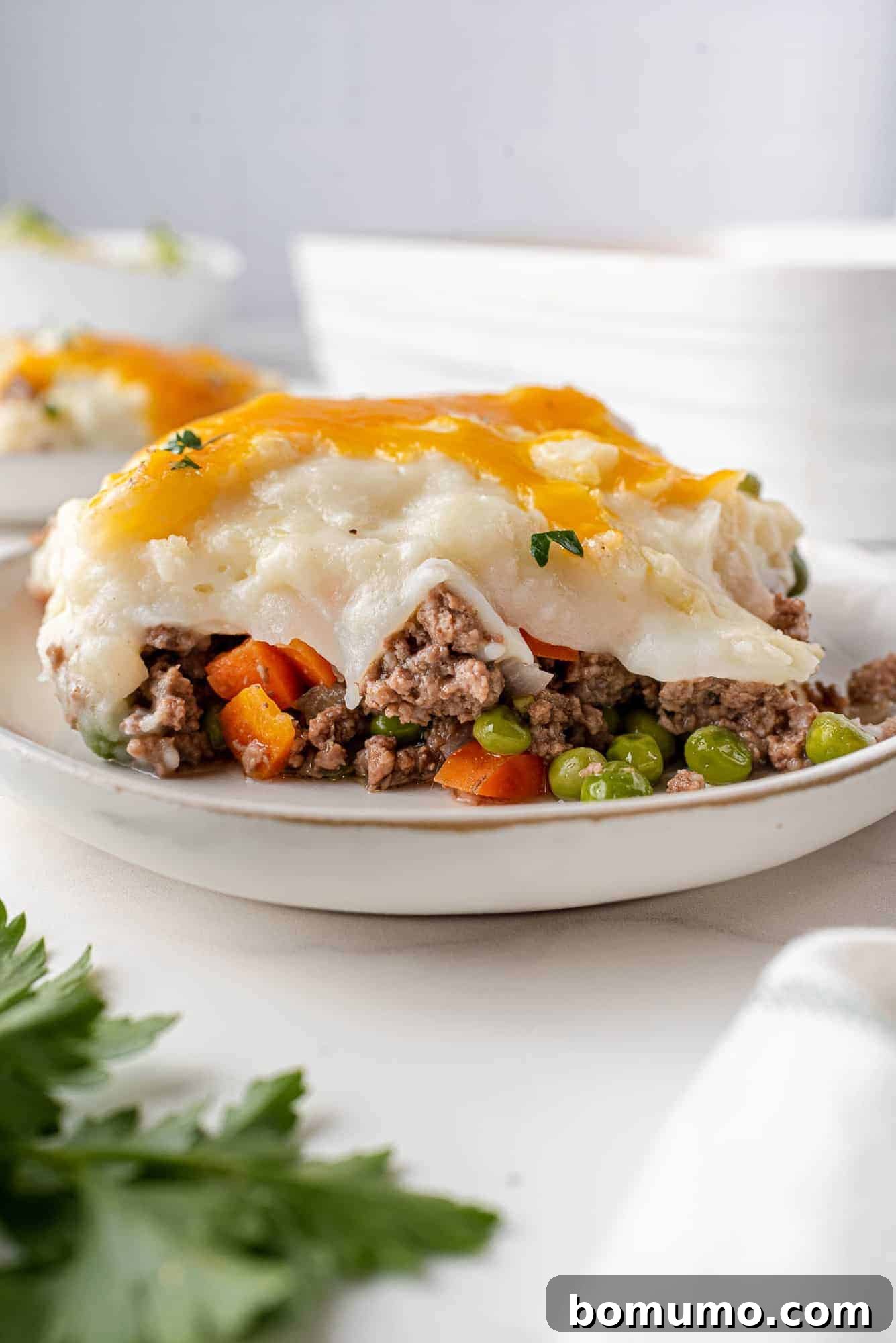 A perfectly portioned slice of shepherd's pie on a pristine white plate, garnished with fresh parsley. The image highlights the beautiful layers: a rich, savory lamb and vegetable base topped with a golden-brown, creamy mashed potato and sharp cheddar cheese crust.