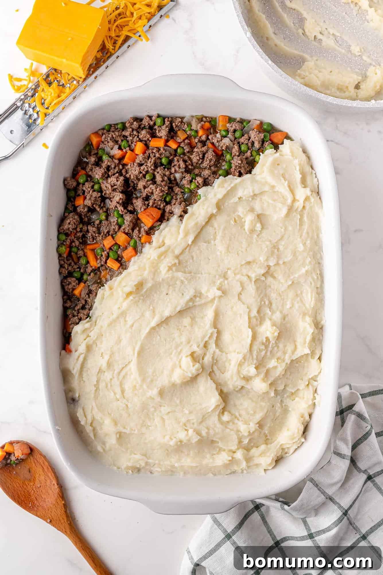 Mashed potatoes being carefully and evenly spread over the savory ground lamb and vegetable filling in a casserole dish, creating a smooth and complete top layer.