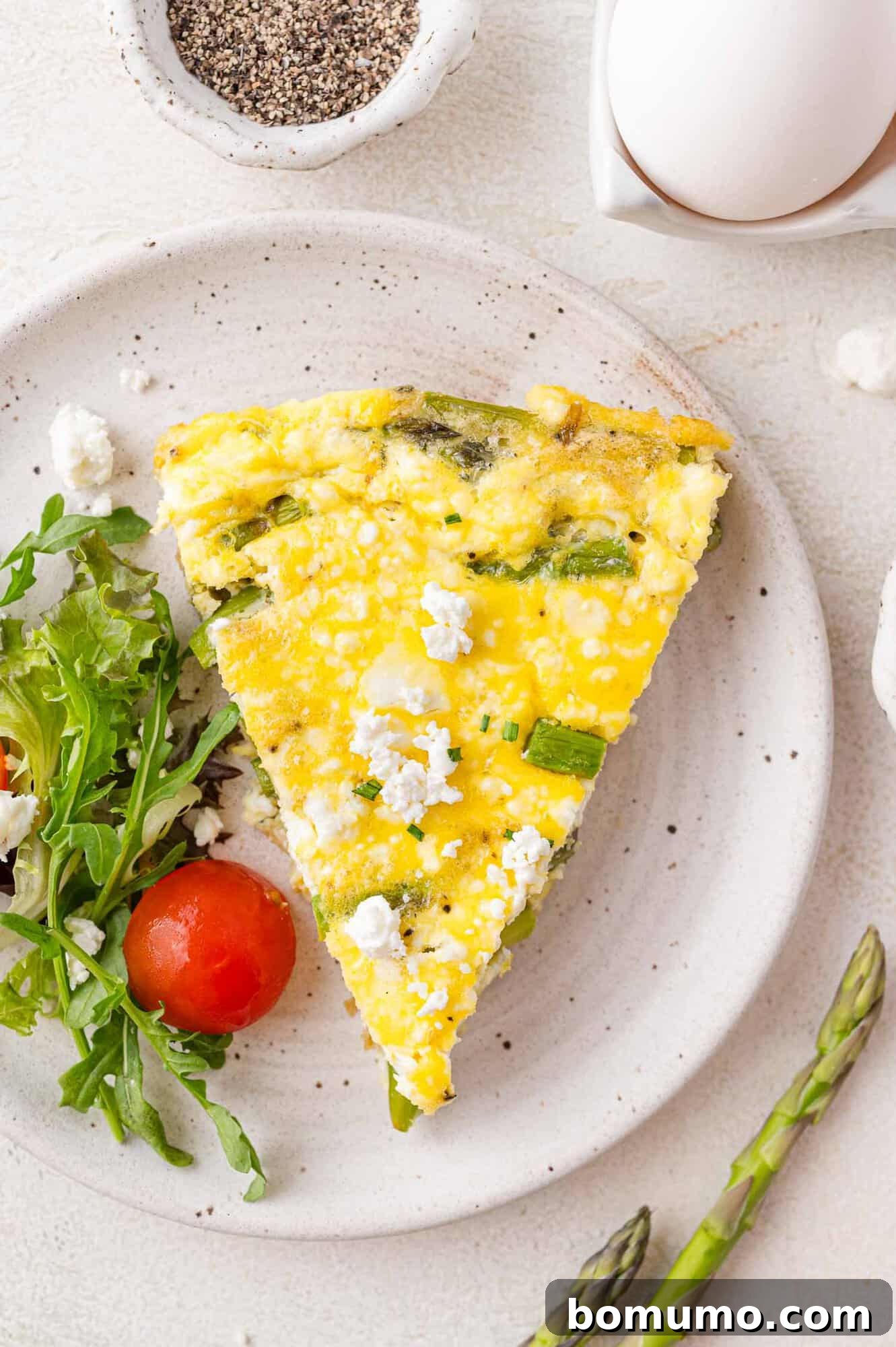 A vibrant slice of asparagus frittata, beautifully plated with crumbled goat cheese, fresh arugula, and a ripe cherry tomato, surrounded by fresh asparagus spears and eggs.