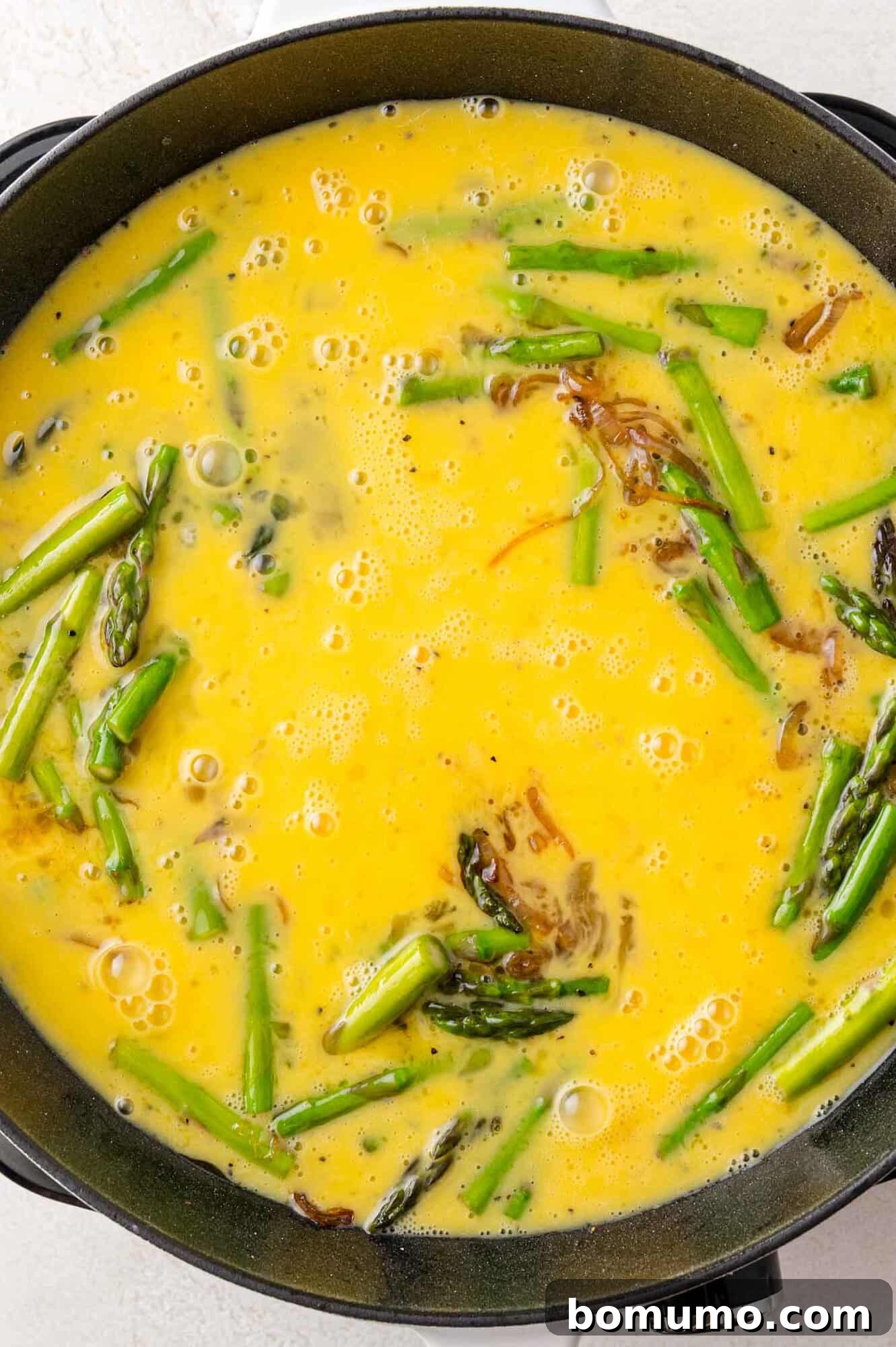 A frying pan filled with a mixture of gently scrambled eggs, vibrant green asparagus, and tender shallots, nearing the point of perfection.