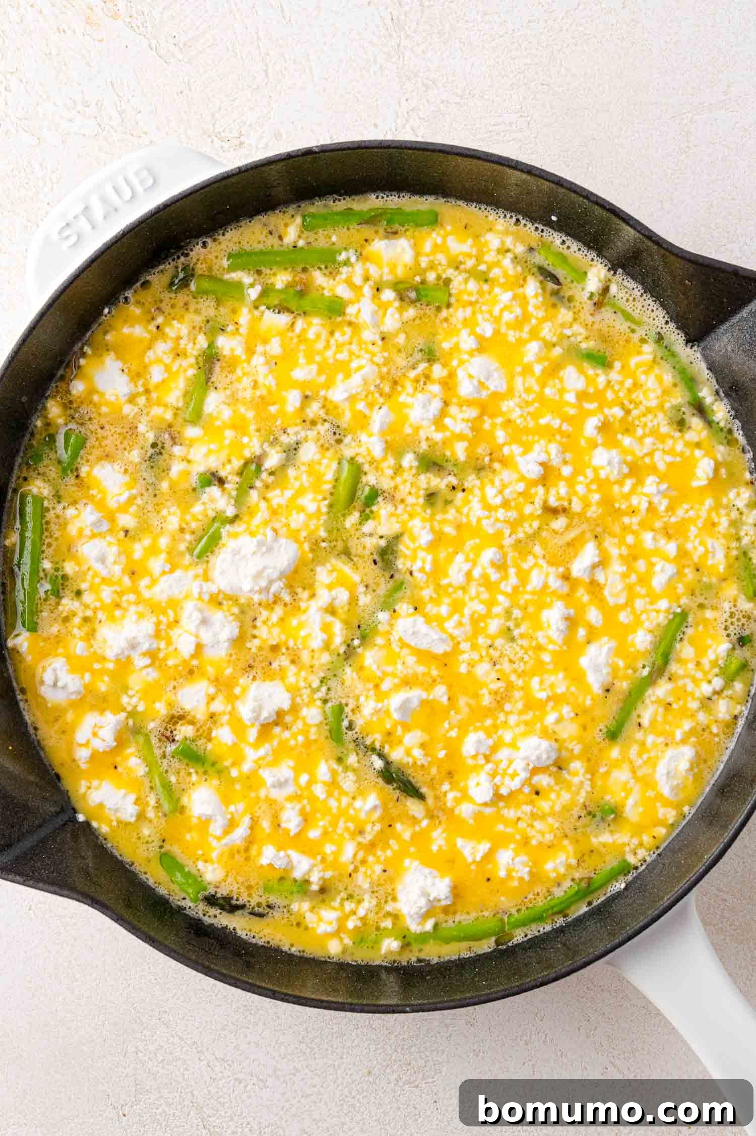 An oven-safe skillet holds a vibrant mixture of beaten eggs, asparagus pieces, and crumbled feta cheese, ready for its final bake to golden perfection.