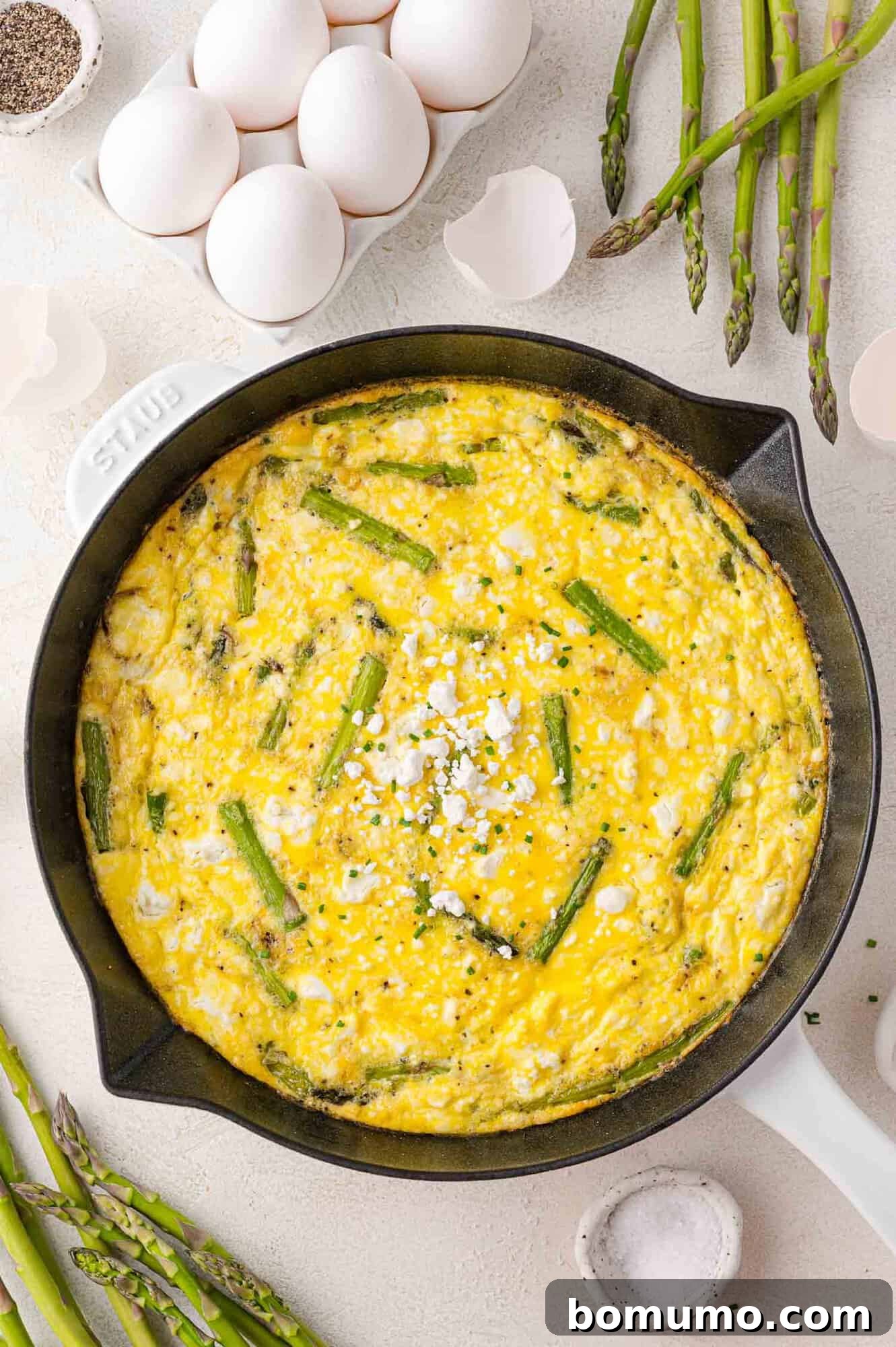 A fresh asparagus frittata in a skillet, beautifully set and golden, surrounded by raw eggs, eggshells, and asparagus spears, ready for serving.