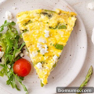 A beautiful slice of vegetarian frittata featuring fresh asparagus and creamy goat cheese on a plate, complemented by a garnish of mixed greens and a cherry tomato.