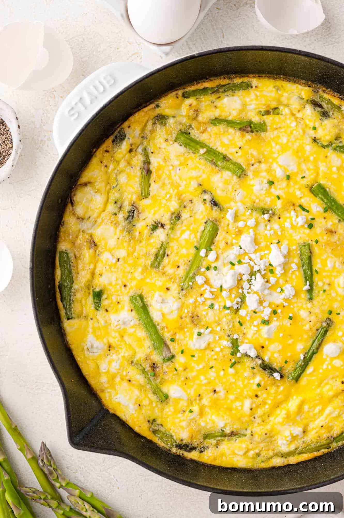 A beautifully cooked asparagus frittata with crumbled goat cheese, nestled in a black oven-safe skillet, surrounded by fresh eggshells and raw asparagus spears.