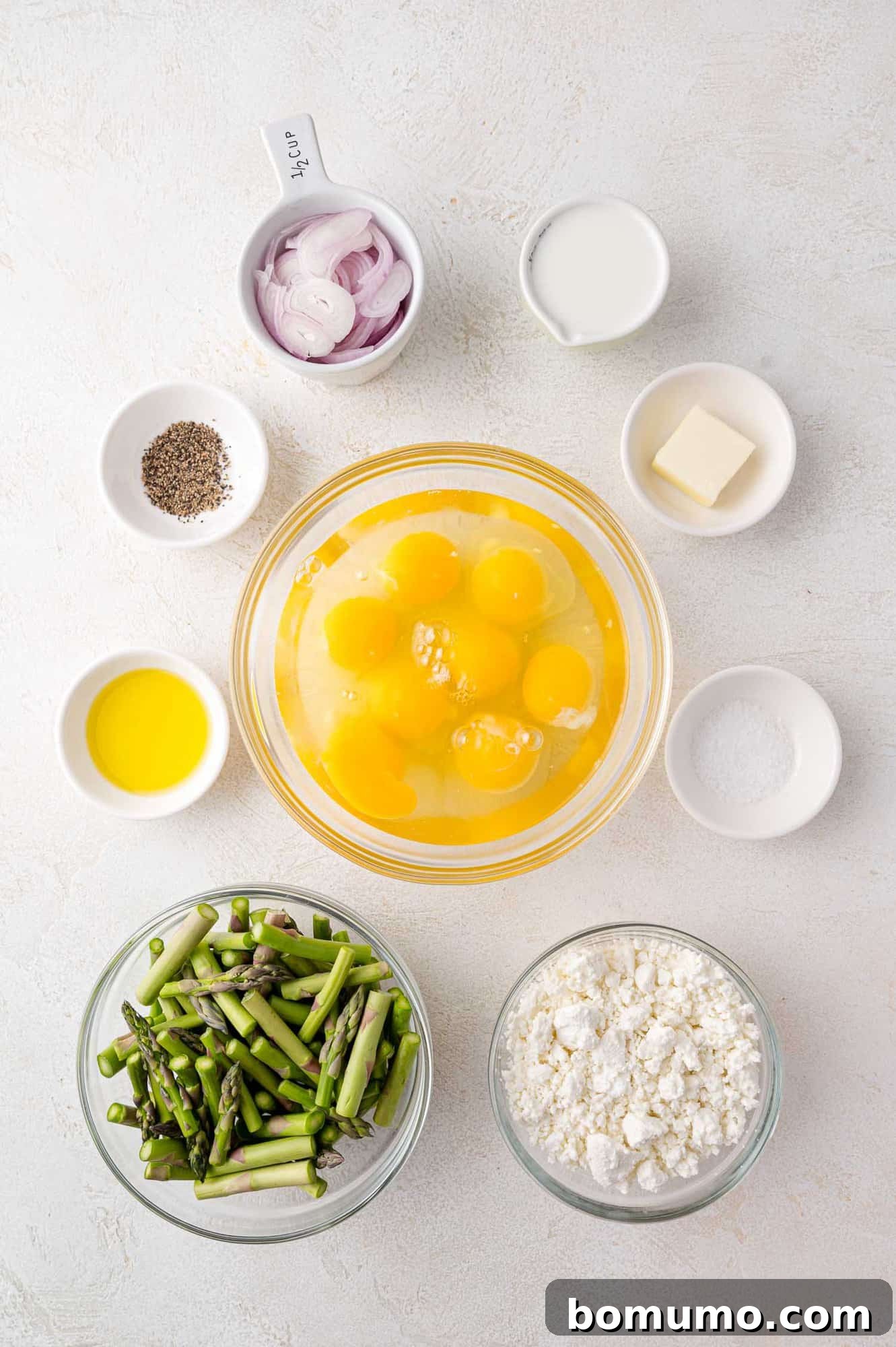 All the fresh ingredients neatly arranged on a countertop: eggs, perfectly chopped asparagus, creamy goat cheese, thinly sliced shallots, a carton of milk, unsalted butter, olive oil, and small bowls of salt and pepper.