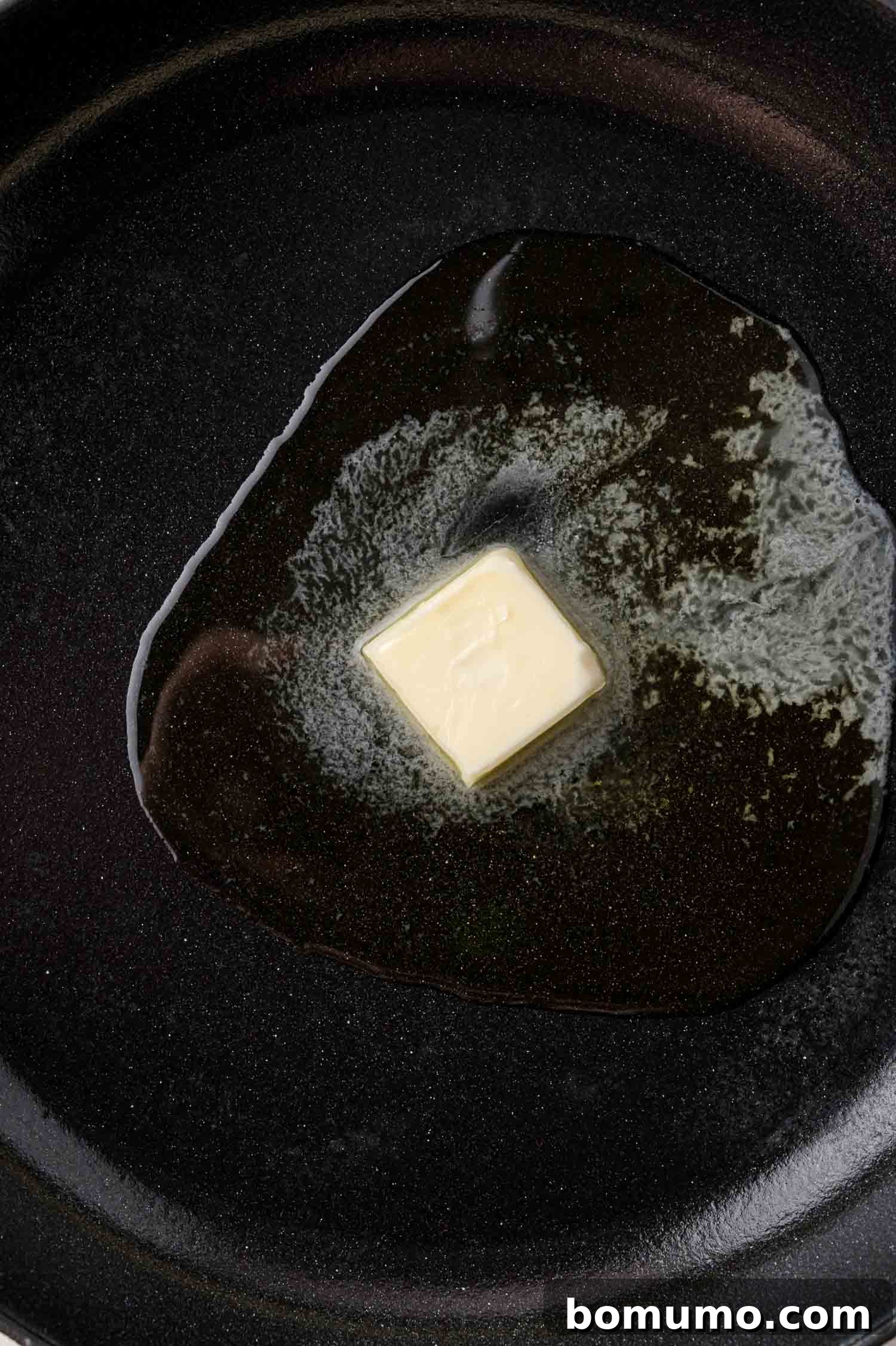 A pat of golden unsalted butter slowly melting in a black nonstick frying pan, signaling the start of a delicious meal.