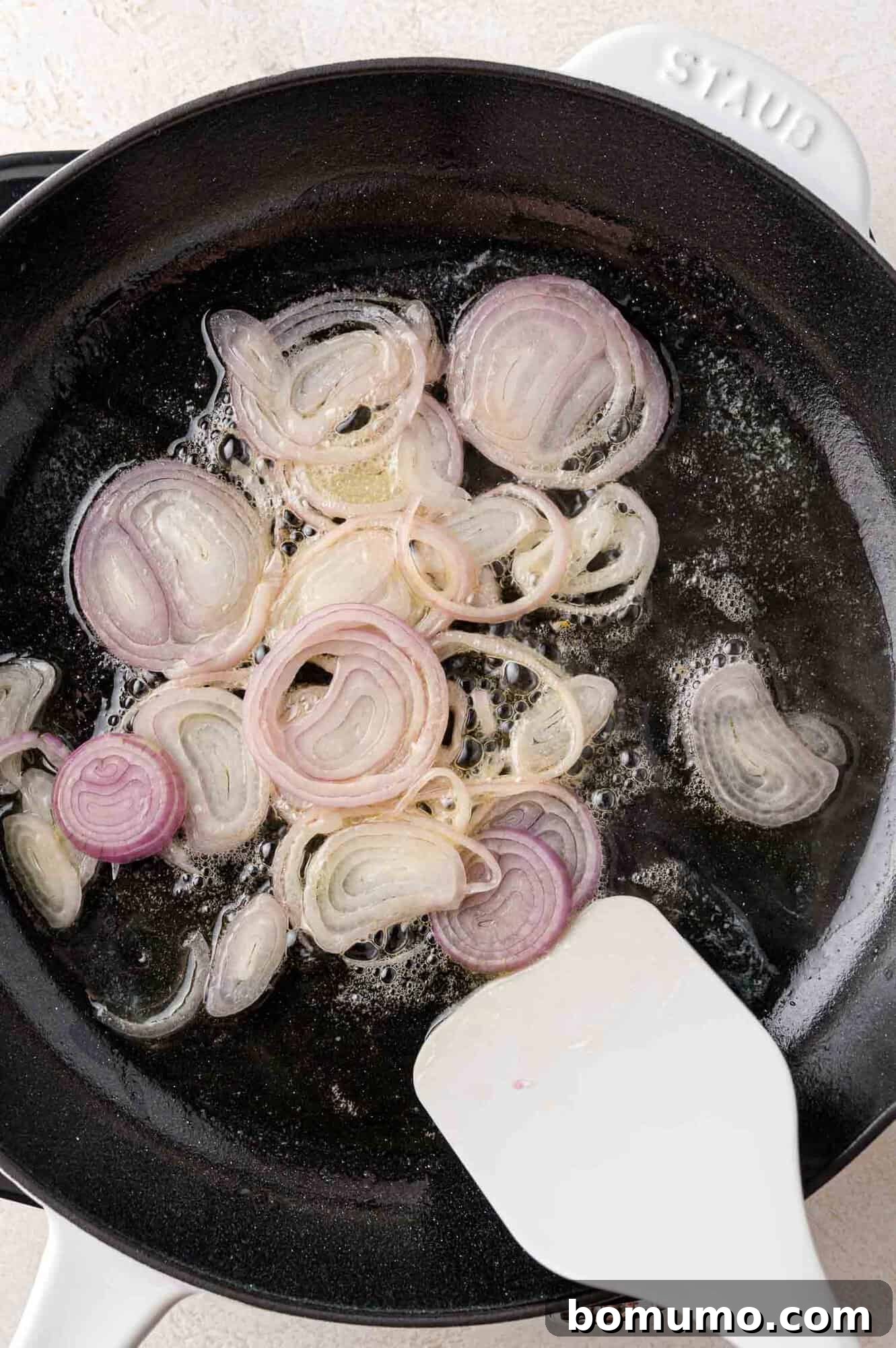 Thinly sliced shallots gently sautéing in a glistening mixture of oil and butter in a black skillet, being stirred with a white spatula.