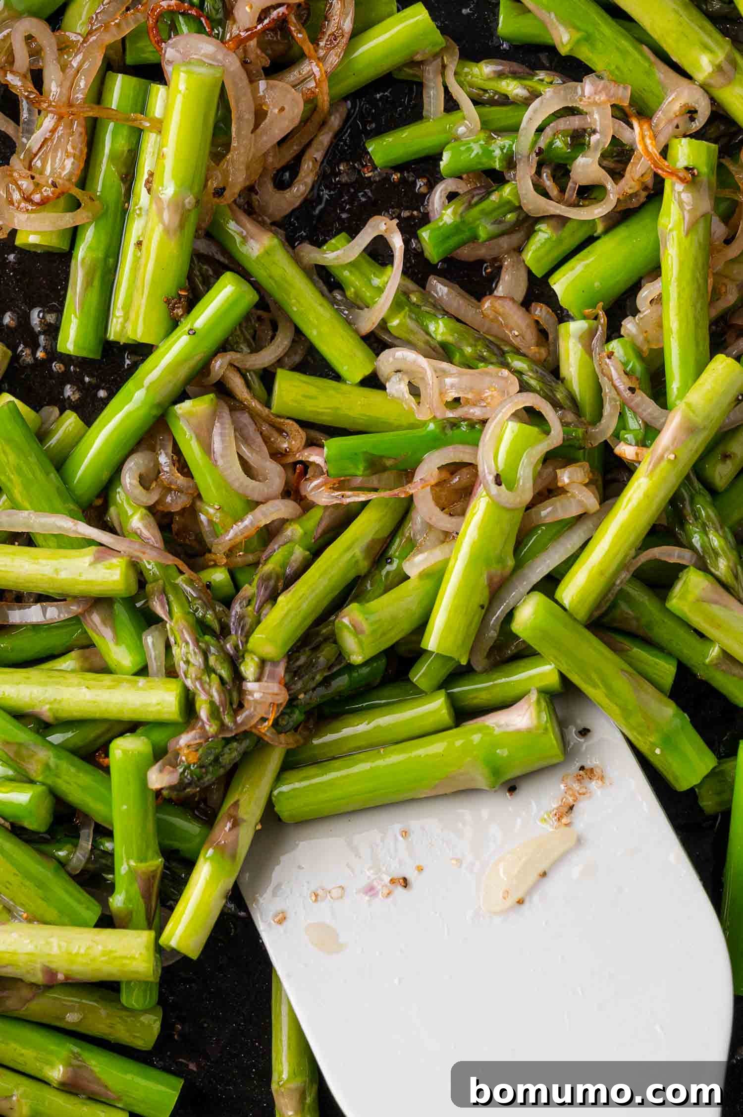 A close-up view of bright green asparagus and translucent shallots expertly sautéing in a pan, being gently tossed with a white spatula.