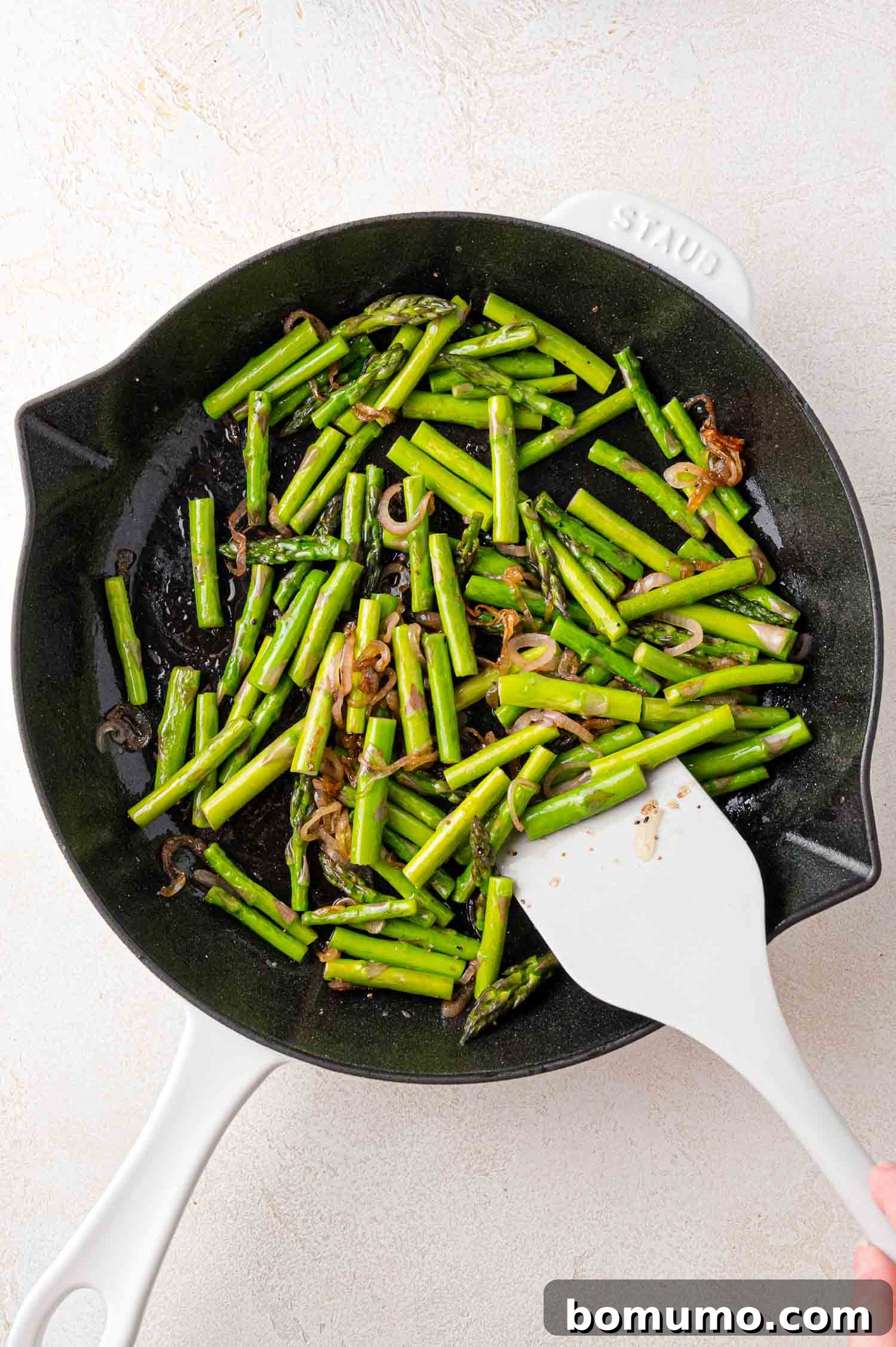 Fresh asparagus and thinly sliced shallots harmoniously sautéing in a white skillet, stirred with a white spatula, perfectly prepped for the frittata.