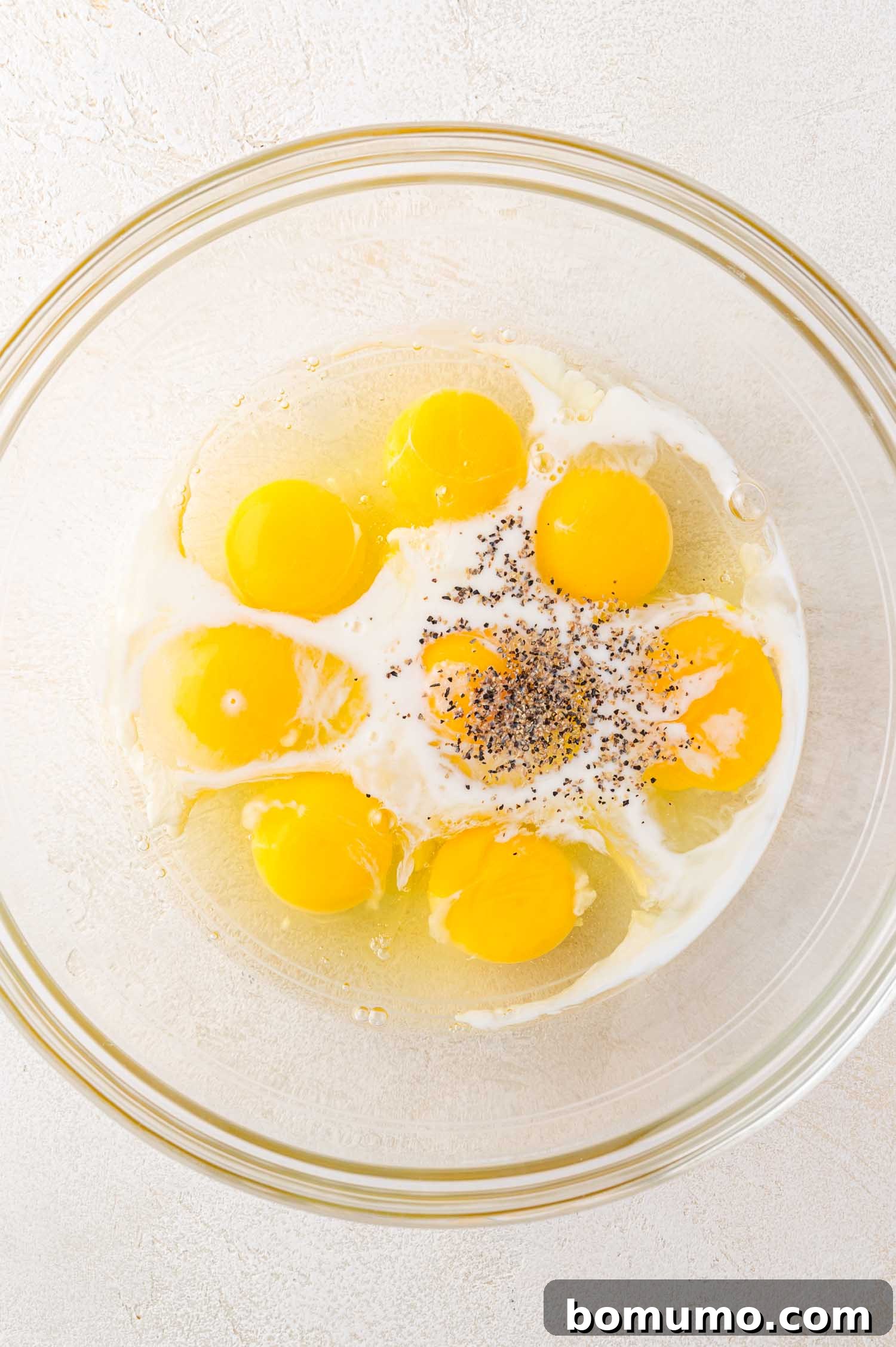 A clear glass bowl on a light-textured surface, holding six raw eggs, a splash of milk, and a sprinkle of freshly ground black pepper, awaiting a whisk.
