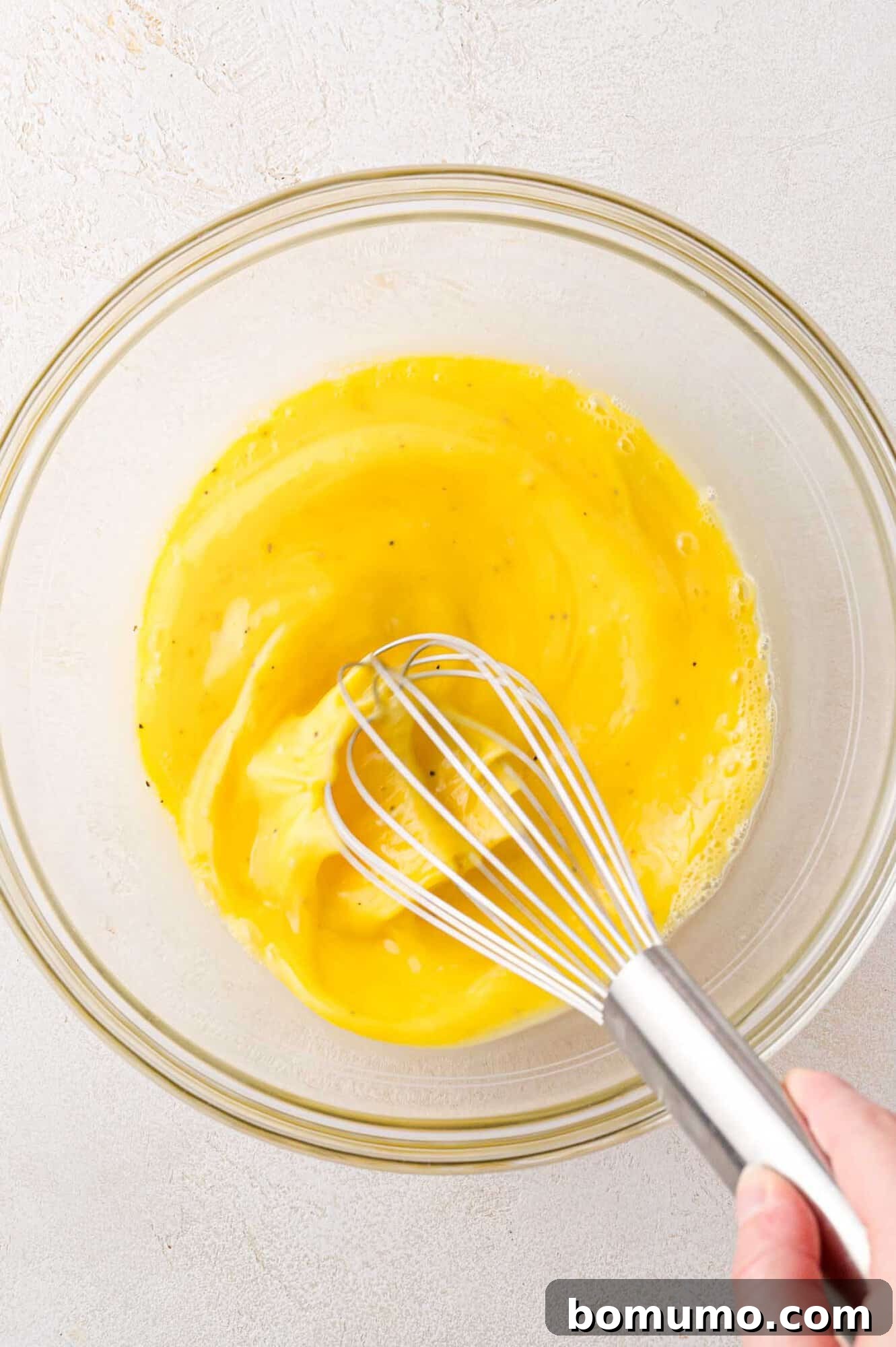 A hand gracefully uses a whisk to combine the golden liquid of eggs and milk in a clear glass bowl, creating a smooth mixture for the frittata.