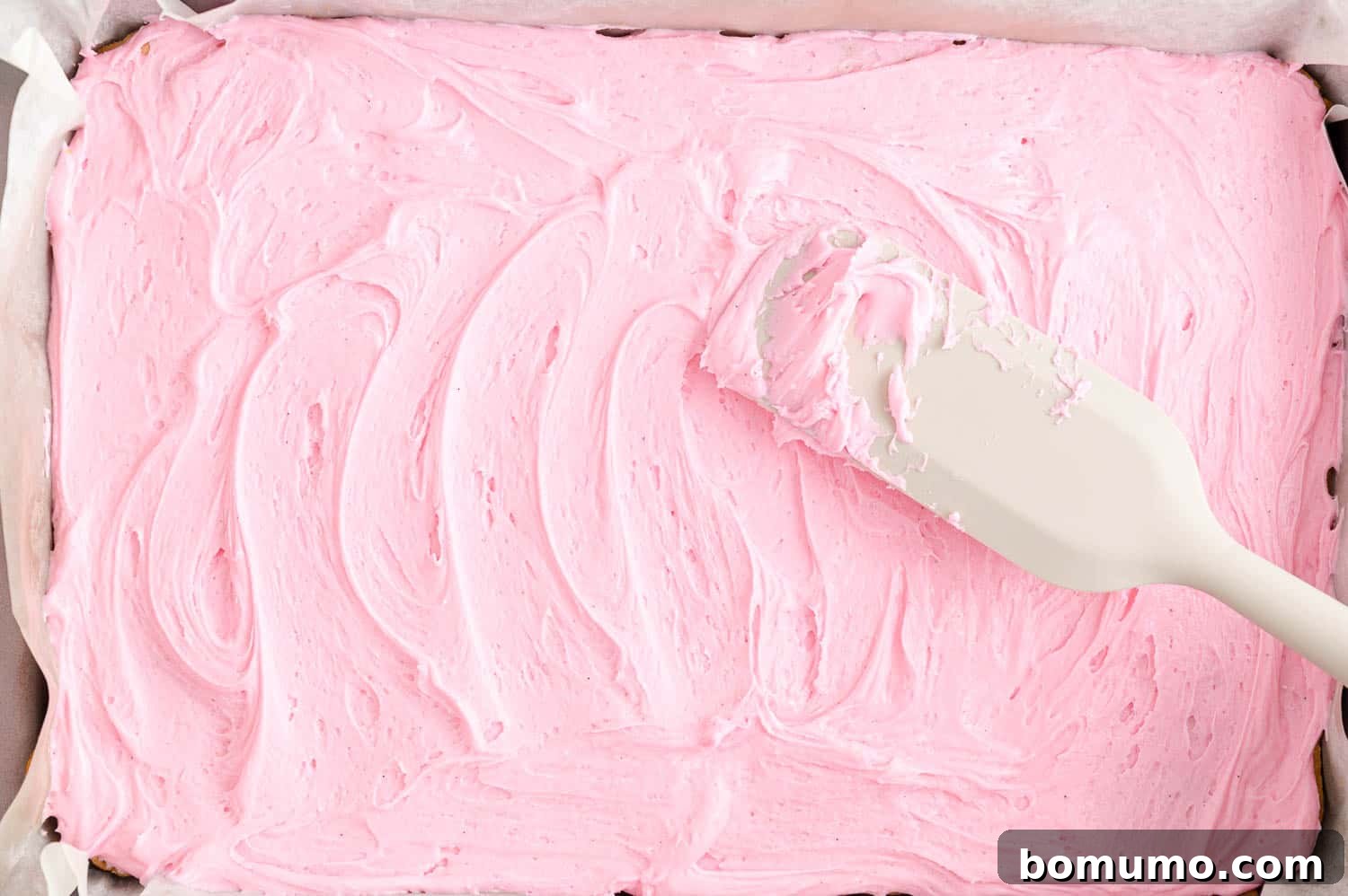 Pink frosting being smoothly spread over the cooled cookie bars, creating an even layer.