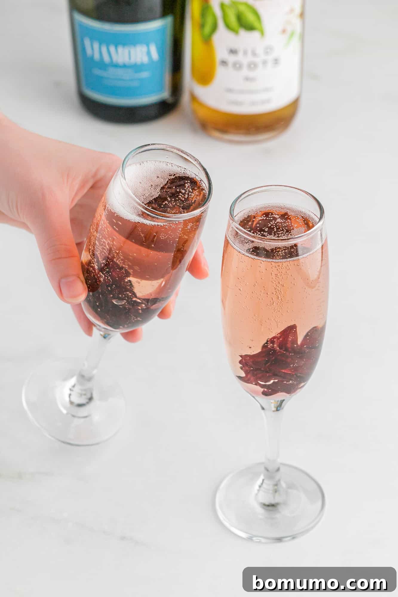 Two champagne flutes, one held in a hand, containing hibiscus mimosas, showcasing their vibrant color.