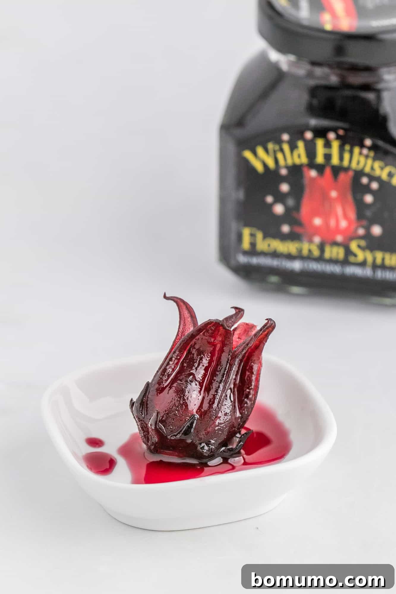 A single hibiscus flower in a small white dish, with the jar in the background, illustrating preparation.