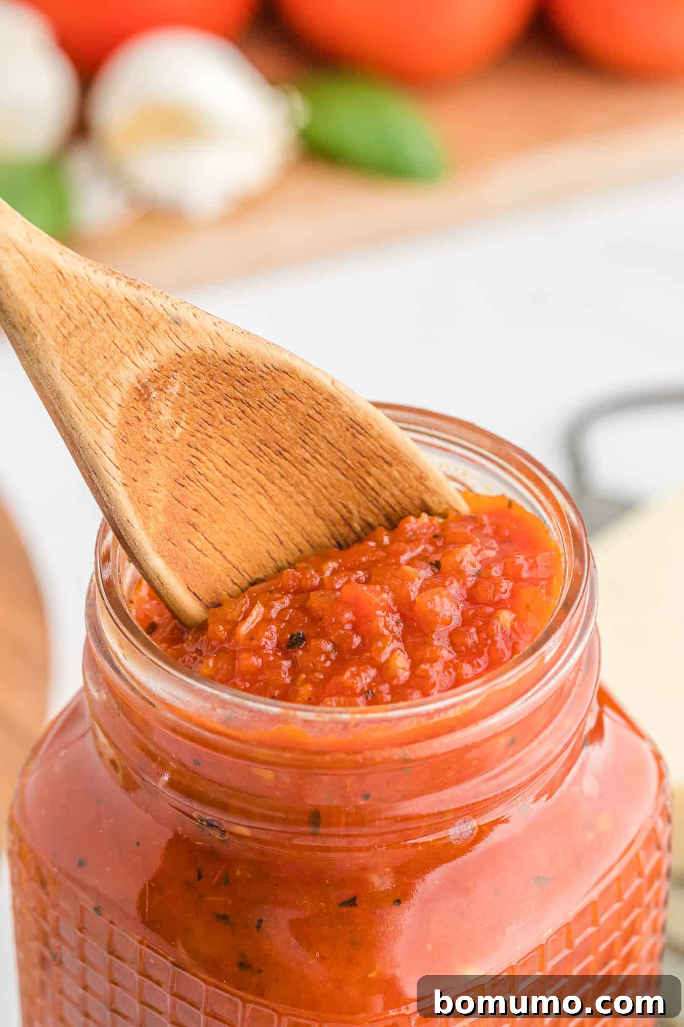 Close-up of a spoon scooping thick, vibrant red pizza sauce from a glass jar, highlighting its rich texture.
