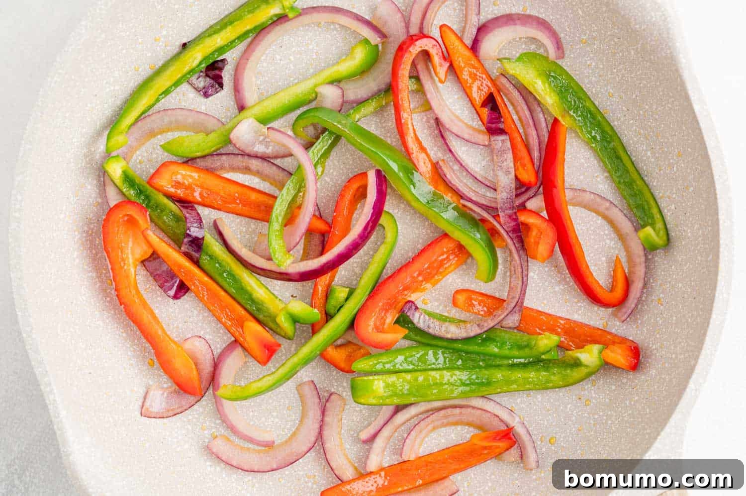 Sautéed peppers and onions sizzling in a white frying pan, ready for the chicken stuffing.