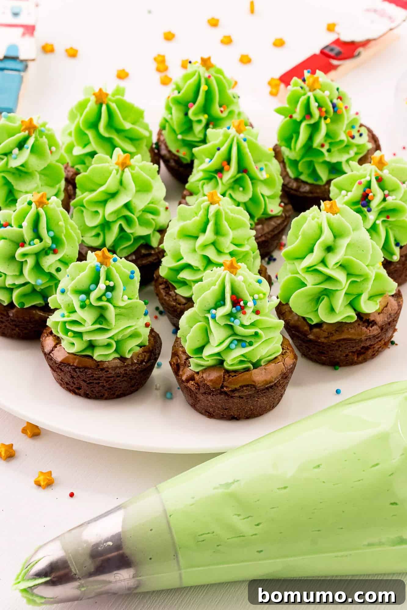 Festive Christmas tree brownies displayed next to a pastry piping bag filled with green frosting, ready for decoration.