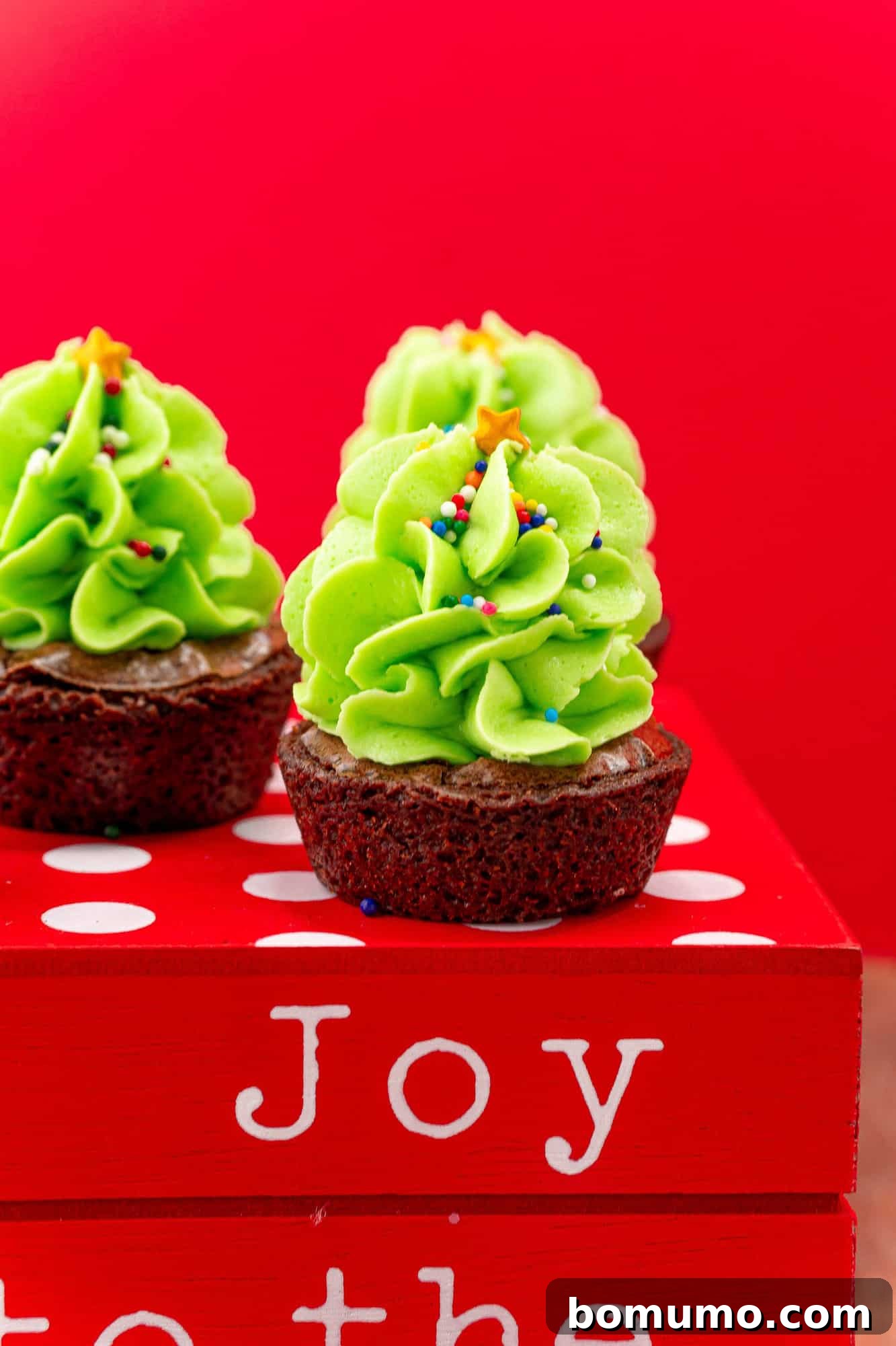 A single, perfectly crafted mini Christmas tree brownie, featuring vibrant green frosting and festive sprinkles, set against a rich red background.