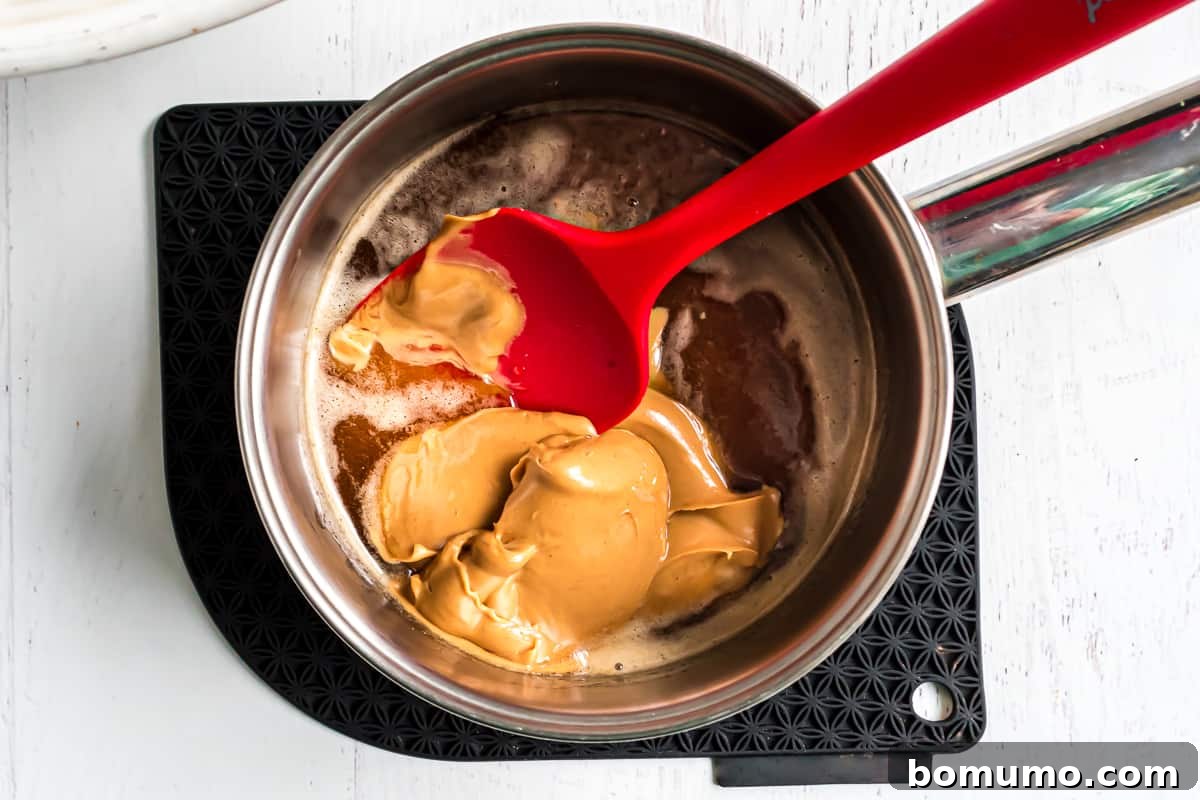 Creamy peanut butter being stirred into the hot sugar and honey mixture in a saucepan.