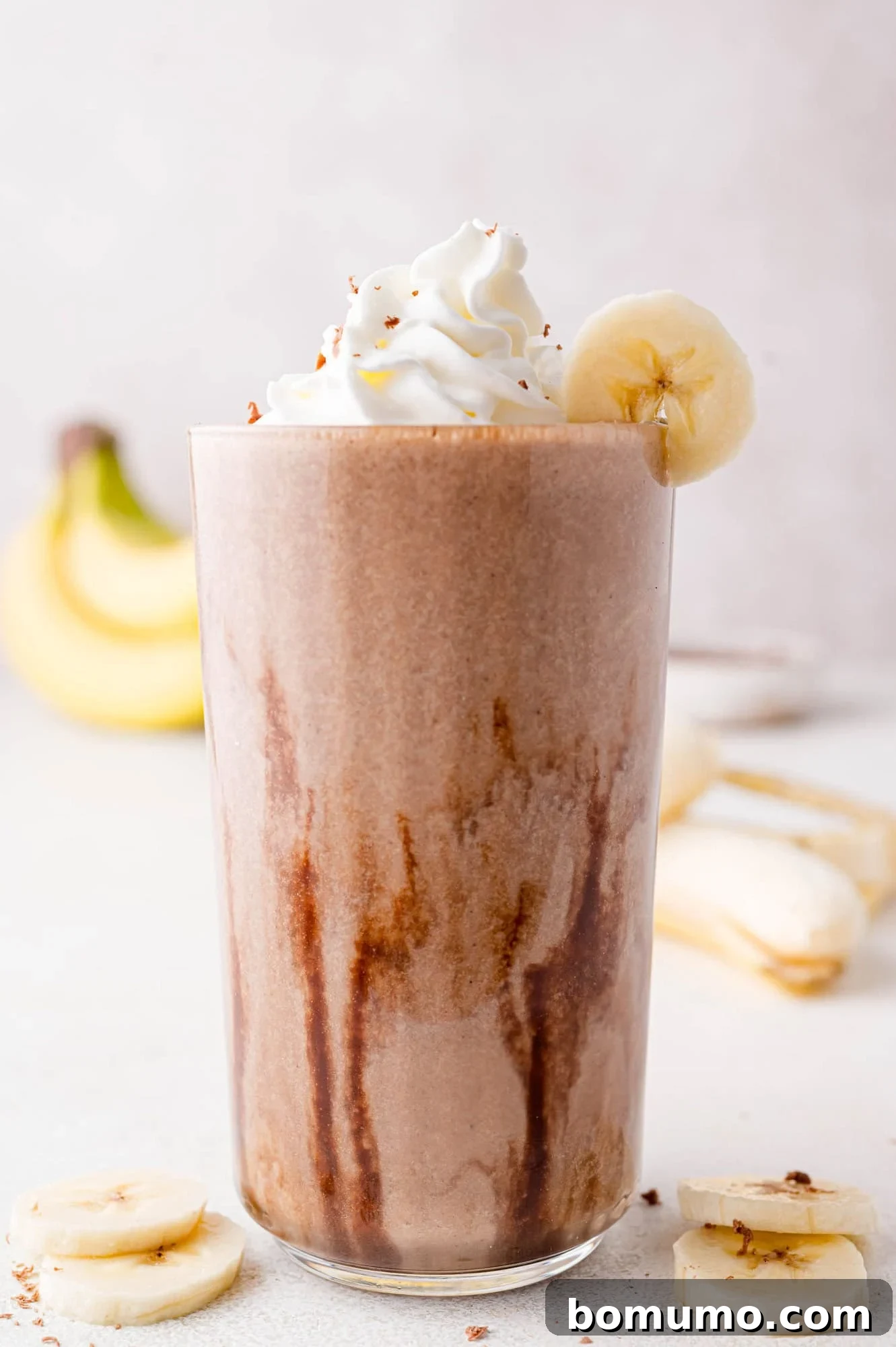 A tall glass of homemade chocolate banana milkshake, crowned with fluffy whipped cream and a fresh banana slice, with ripe bananas artfully blurred in the background.
