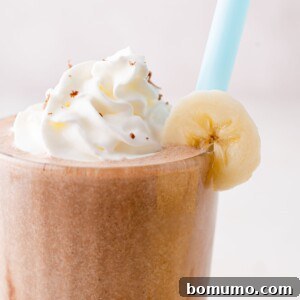 A creamy chocolate banana milkshake, topped with a generous swirl of whipped cream and a delicate banana slice on the rim, complete with a blue straw.