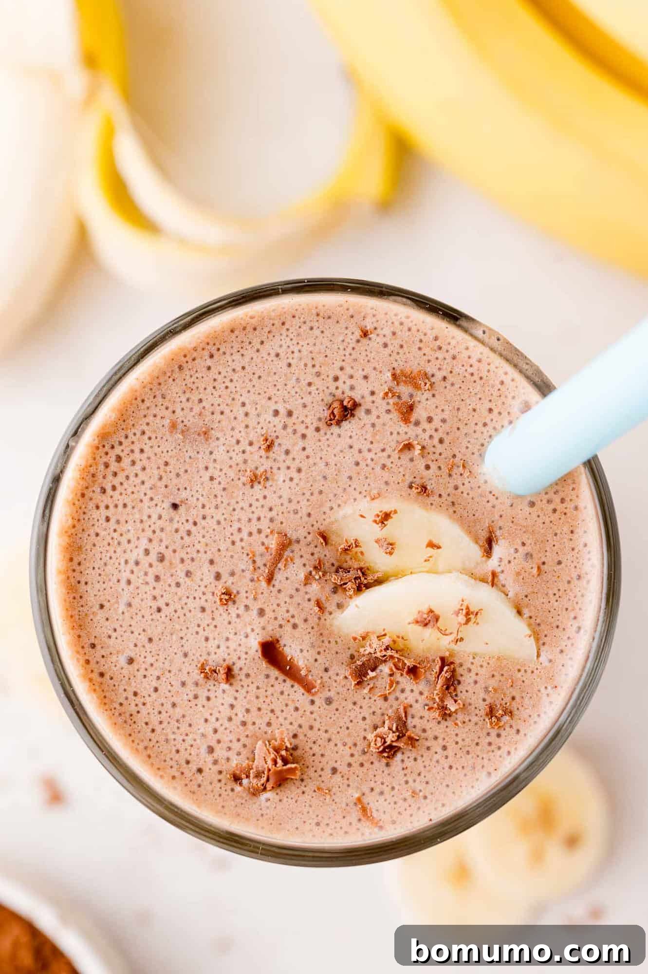 A vibrant, refreshing glass of chocolate banana milkshake, garnished with delicate chocolate shavings and fresh banana slices, served with a blue straw for sipping enjoyment.
