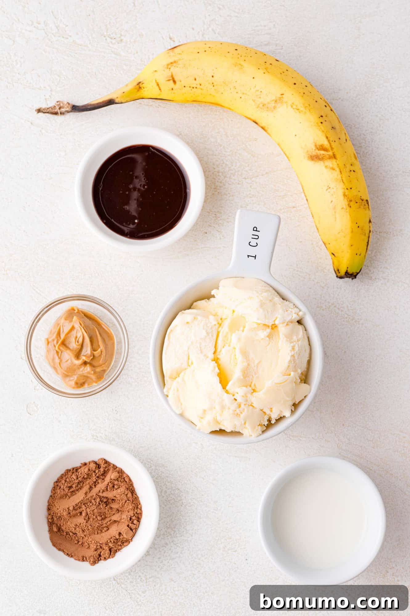 A beautifully arranged selection of essential ingredients for a chocolate banana milkshake: a ripe banana, scoops of vanilla ice cream, unsweetened cocoa powder, creamy peanut butter, rich chocolate sauce, and fresh milk, all set on a clean white surface.