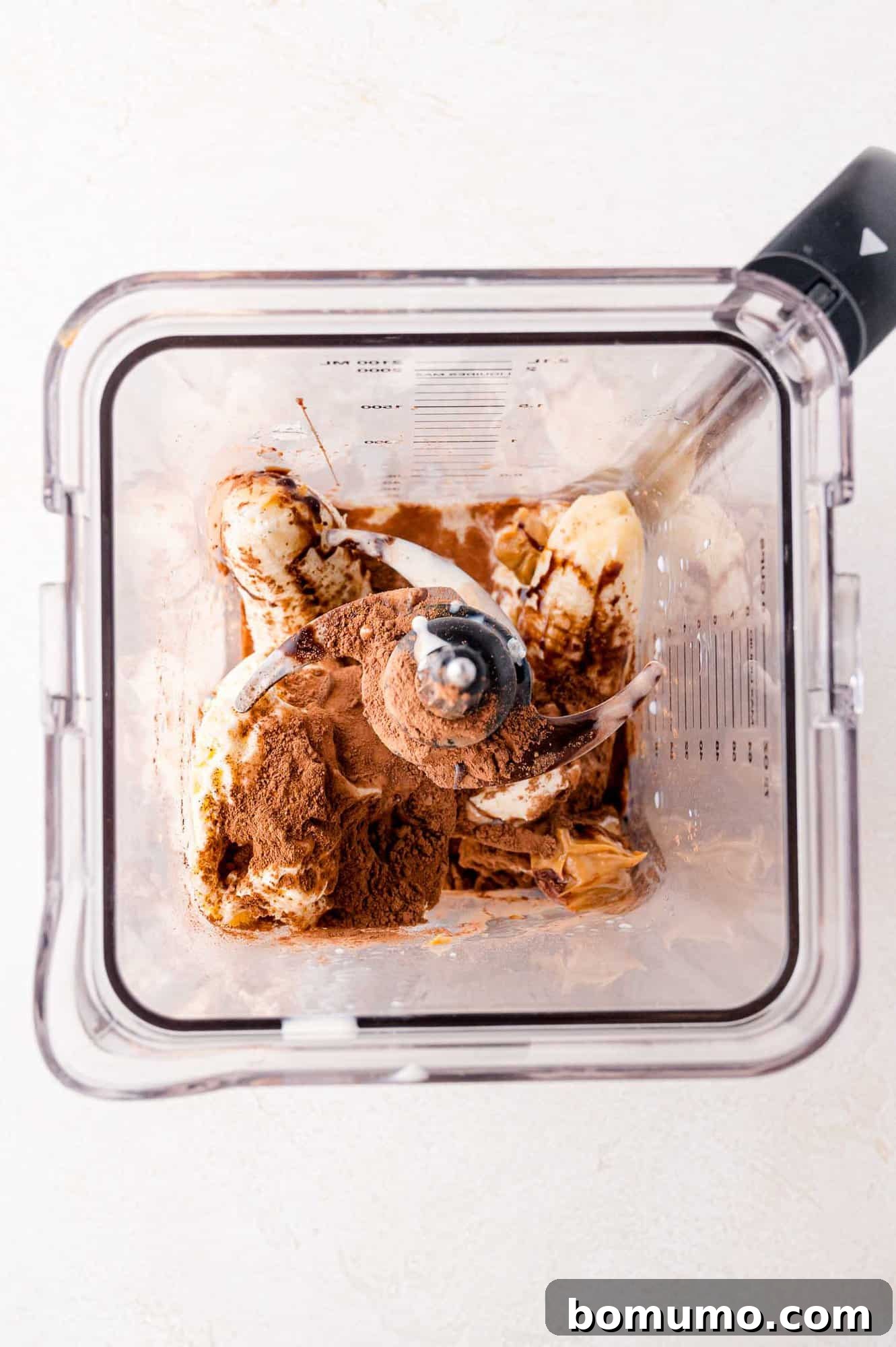 An overhead view inside a blender, revealing neatly sliced banana pieces, a spoonful of rich cocoa powder, and a dollop of creamy peanut butter, ready to be blended.