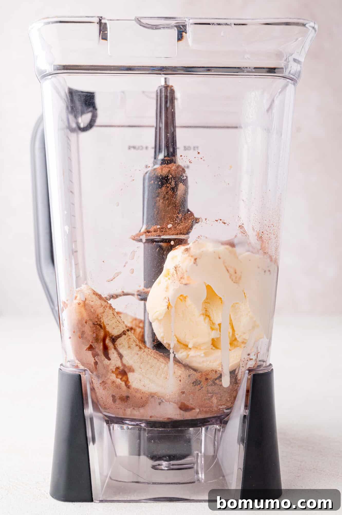 A blender container filled with generous scoops of vanilla ice cream, a ripe banana, and a sprinkle of chocolate powder, poised for blending into a delicious milkshake.