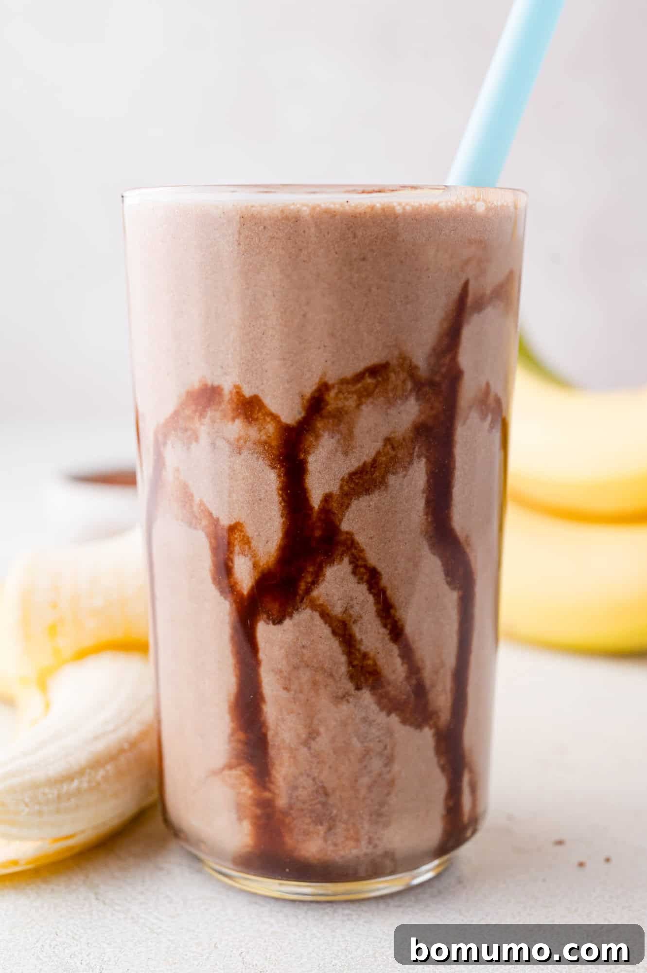 A delightful glass of chocolate banana smoothie, elegantly drizzled with chocolate syrup and accompanied by a blue straw, with a backdrop of fresh bananas.