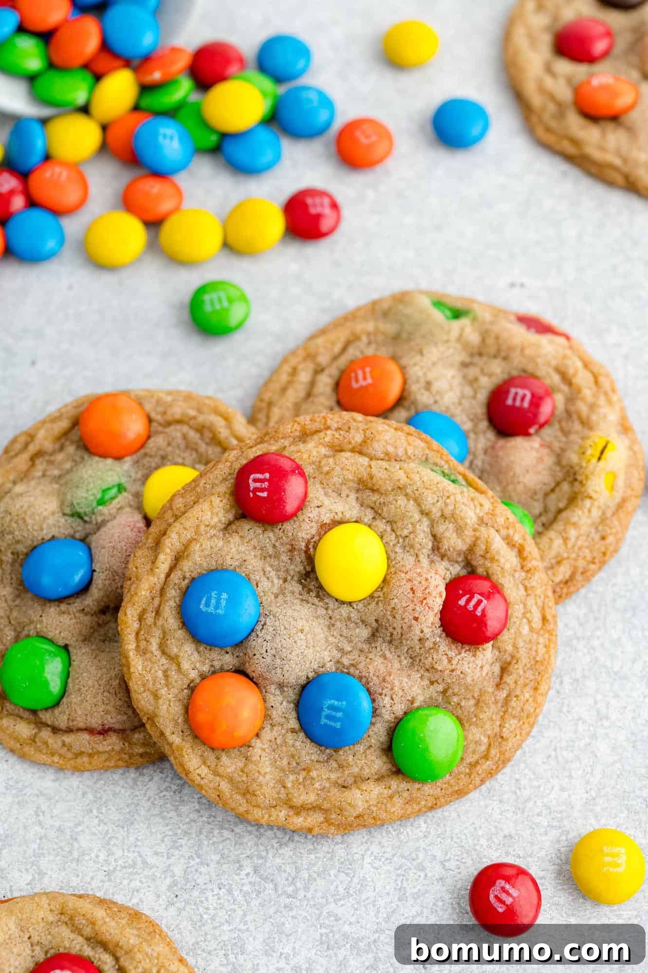 Ultimate M M Cookies 12 A large, inviting pile of M&M cookies, with a bowl of extra M&Ms in the background, hinting at their fresh preparation.