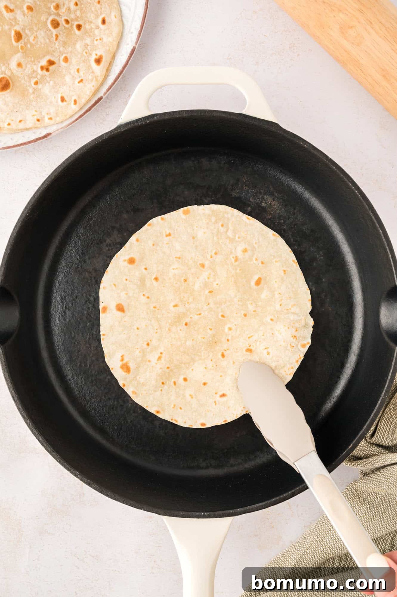 A tortilla being flipped over in a cast iron pan with tongs, revealing light charring.