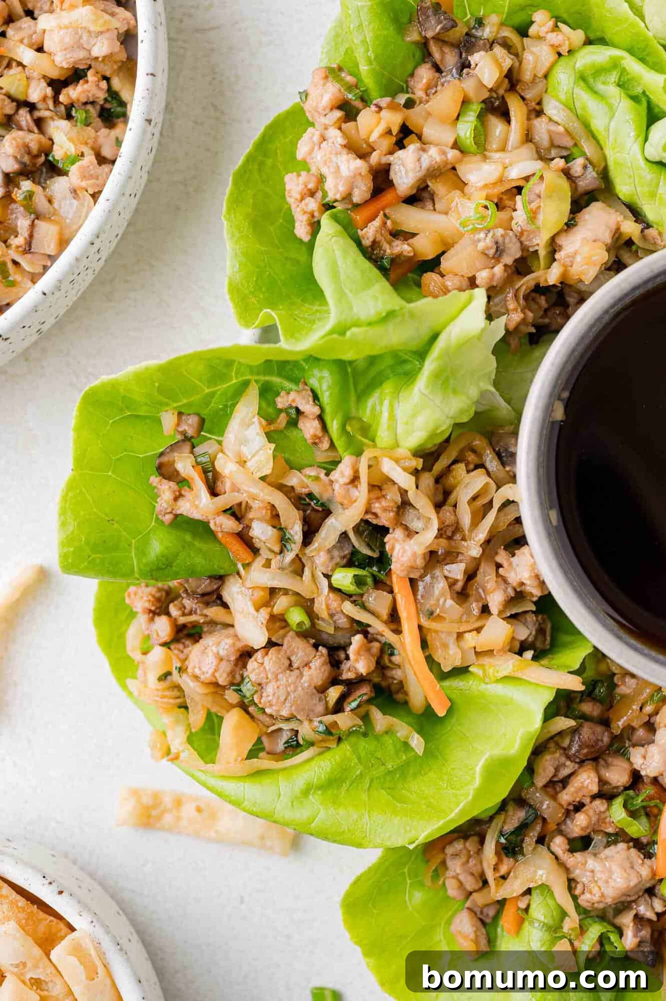 Lettuce wraps filled with cooked ground meat, chopped vegetables, and a bowl of sauce on a light surface.