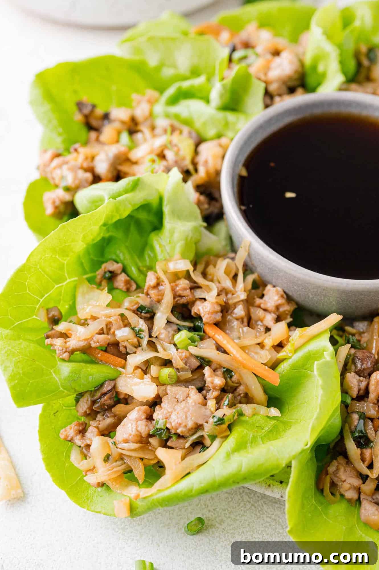 Fresh lettuce wraps filled with minced chicken, vegetables, and herbs are arranged around a small bowl of dipping sauce on a white background.