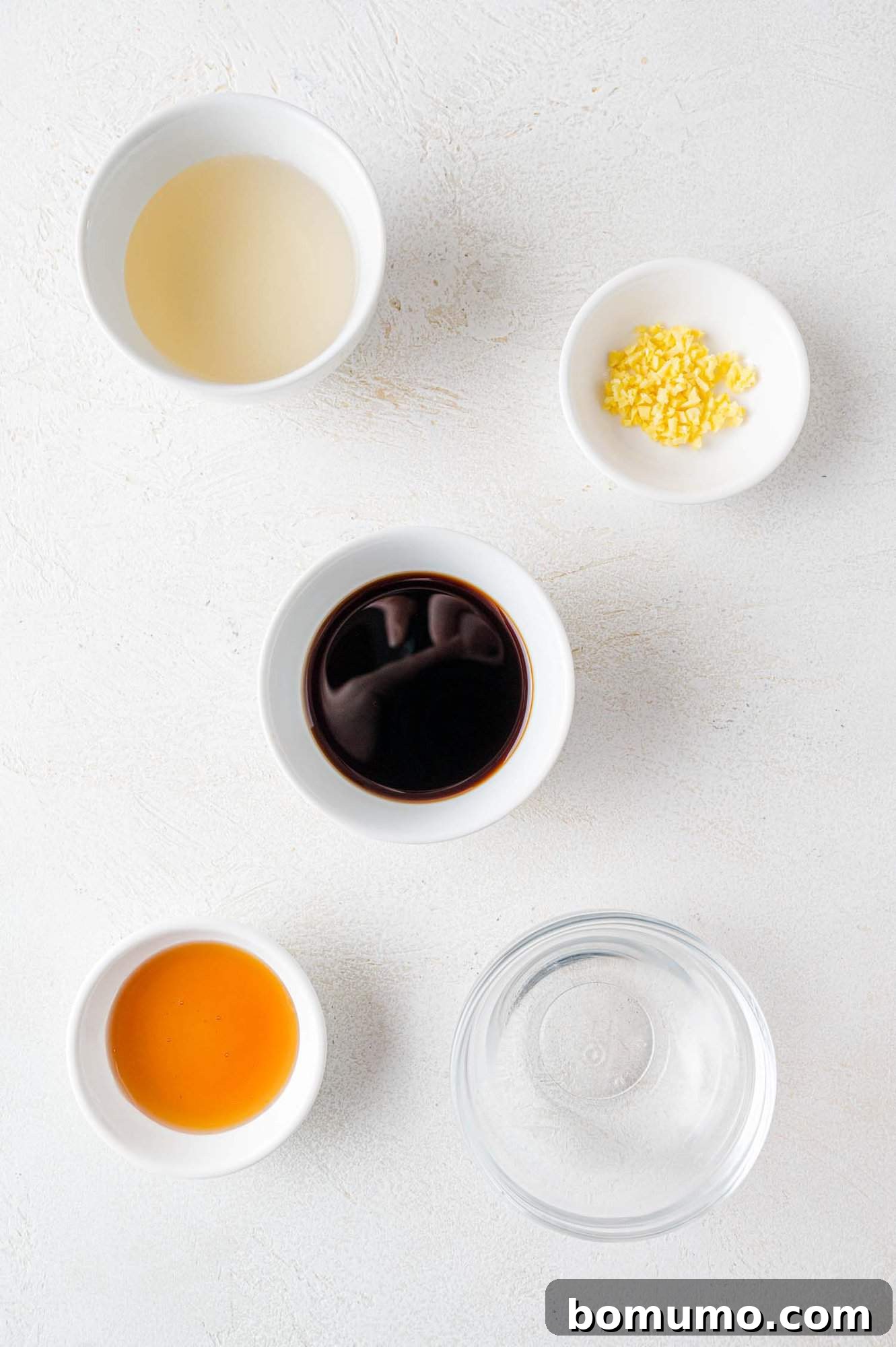 Five small bowls containing various liquid ingredients and minced garlic on a white surface.